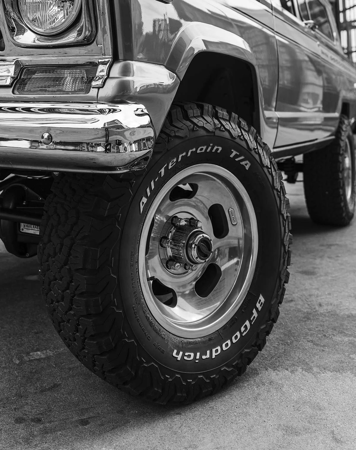 Close-up of a large off-road tire on a vintage off-road vehicle in black and white. The tire has deep treads and white lettering that reads 'BFGoodrich All-Terrain T/A'.
