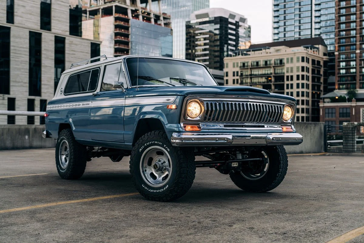 A vintage blue Jeep Wagoneer parked in an urban parking lot with tall buildings in the background.