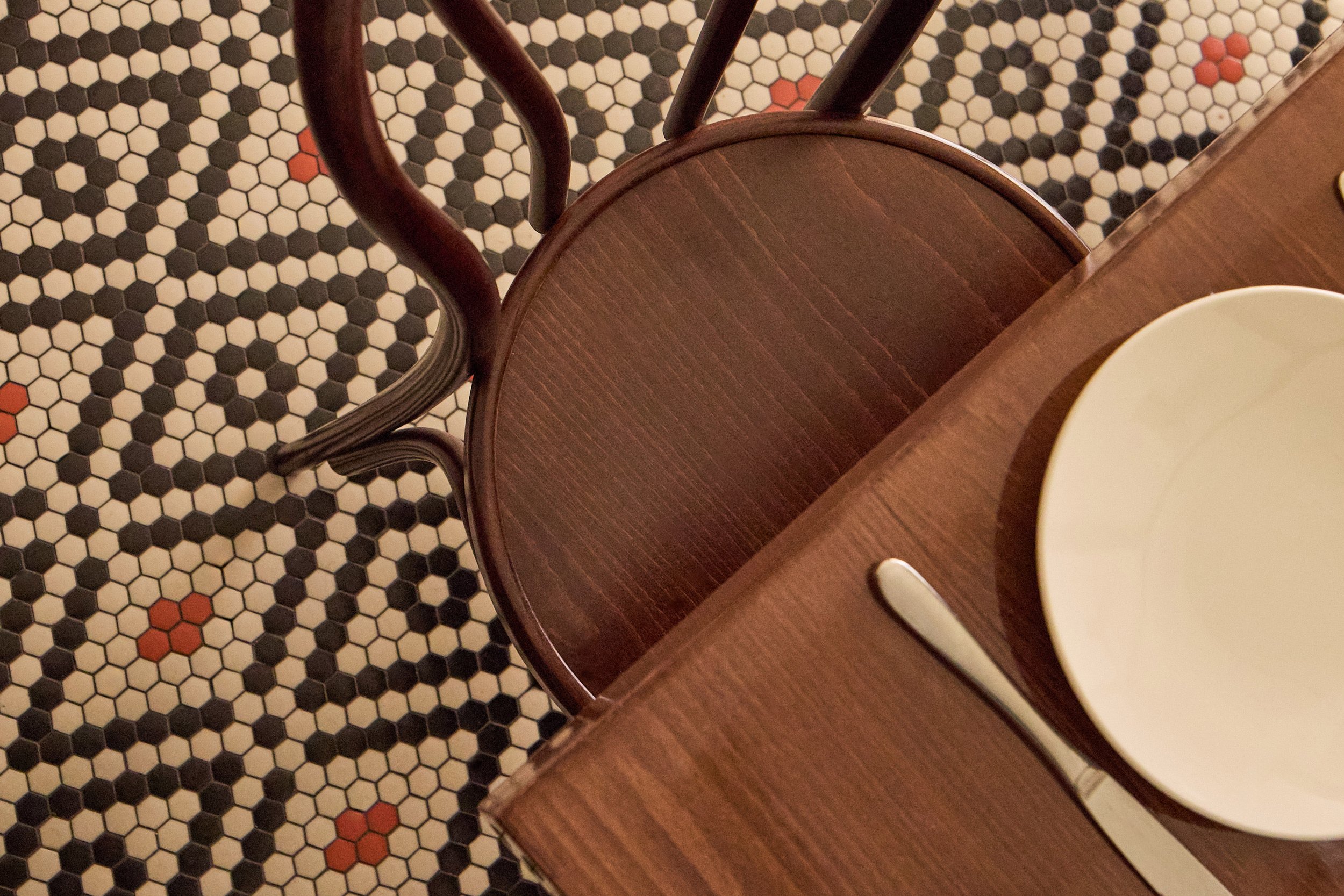 Vista aérea de una silla de madera junto a una mesa en un suelo de mosaico hexagonal blanco, negro y rojo. Sobre la mesa hay un plato blanco y un cuchillo de metal.