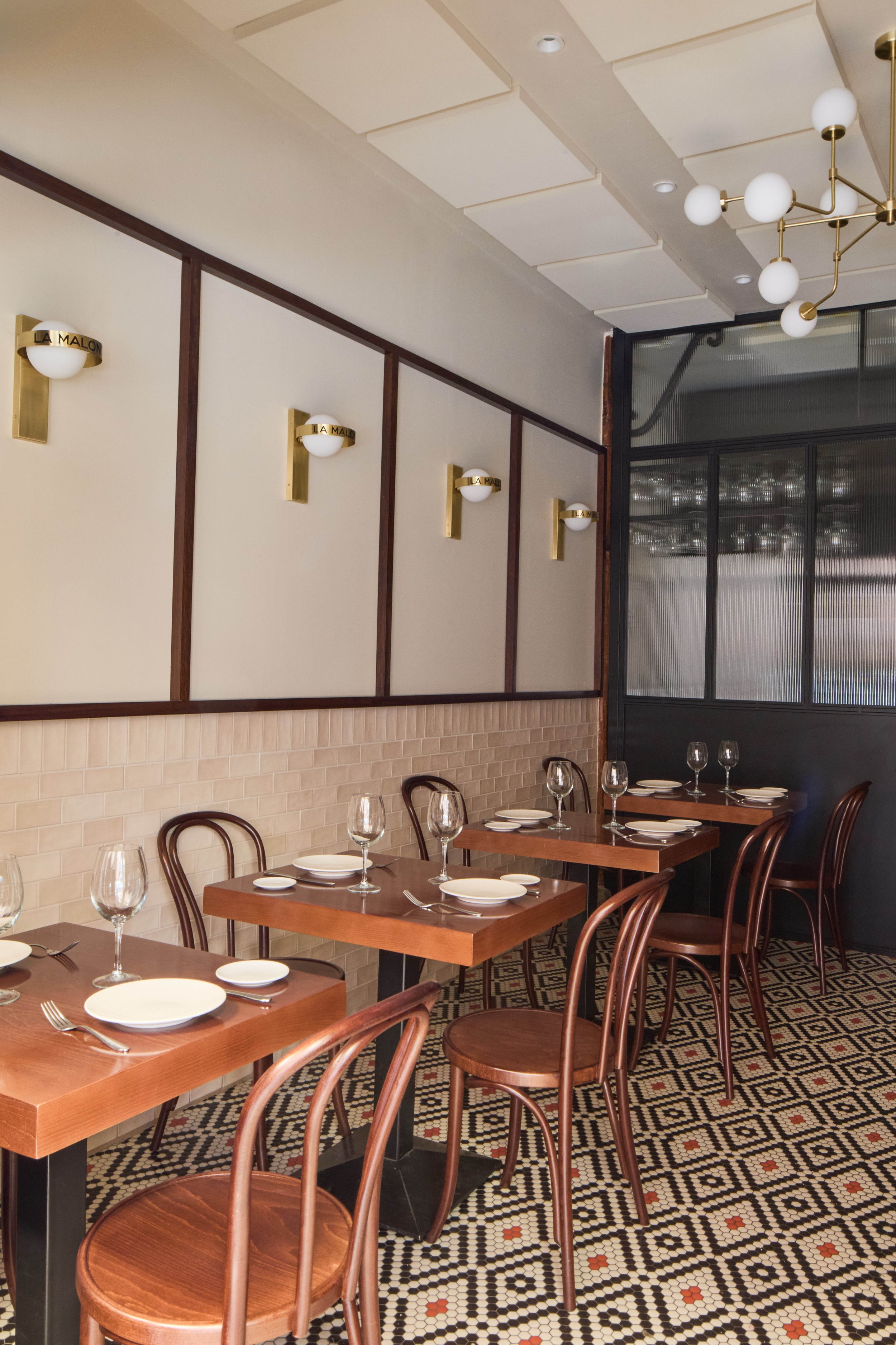 Restaurante con mesas de madera, sillas de diseño clásico, copas de vidrio, platos y cubiertos. Paredes con apliques de luz dorados y techo con lámpara moderna. Suelo con patrón de mosaico.