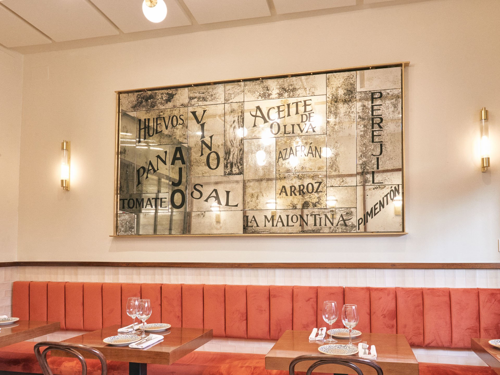 Interior de un restaurante con decoración de paredes en tonos claros y bancos acolchados de color naranja. En la pared se observa un cuadro grande con palabras referentes a ingredientes culinarios como "Huevos", "Vino", "Aceite de Oliva", "Arroz" y "