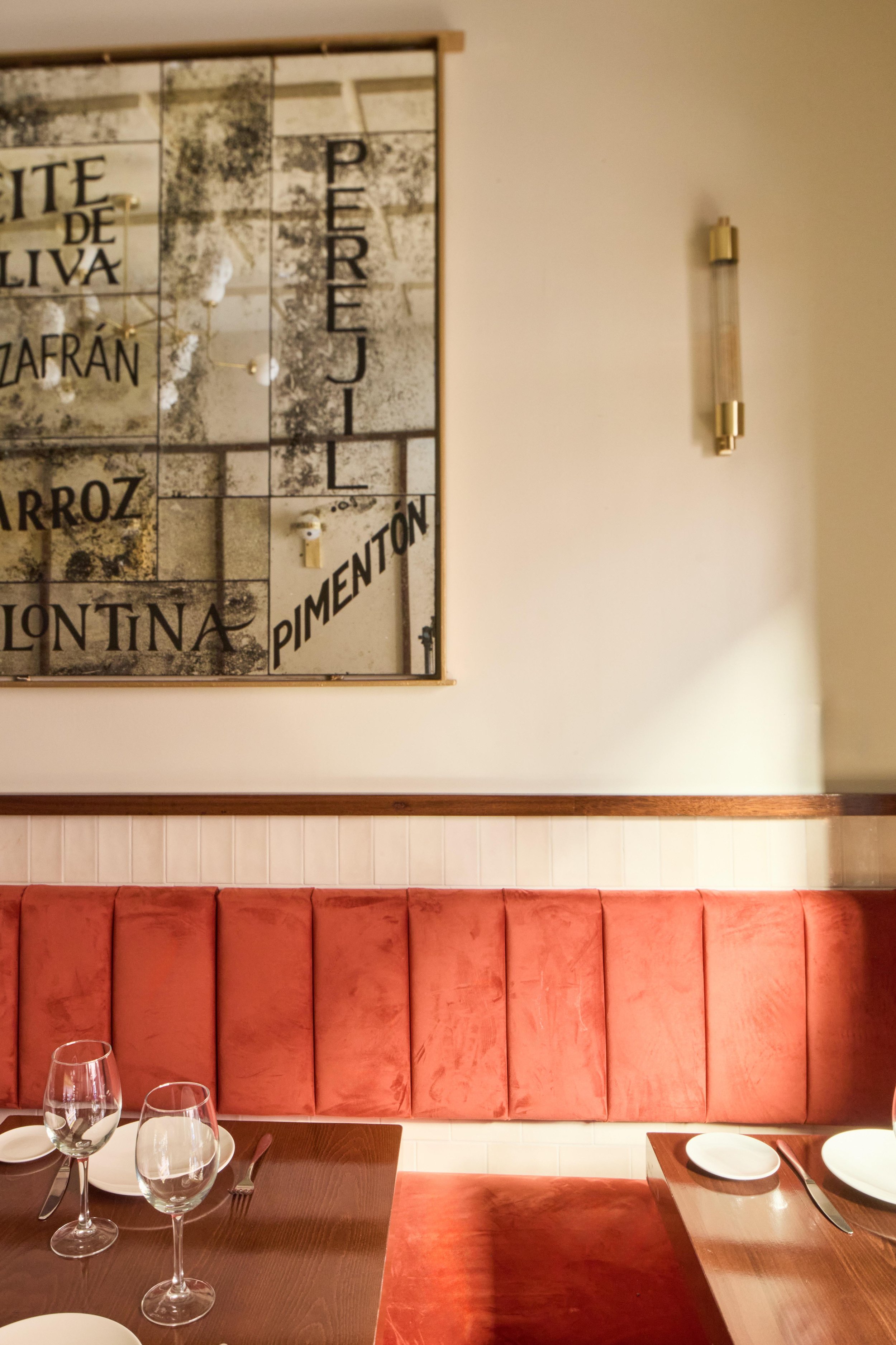 Interior de un restaurante con un sofá rojo, una mesa de madera con copas y platos, y un cartel decorativo en la pared mostrando palabras como 'perejil' y 'pimentón'. Iluminación cálida con una lámpara de pared.