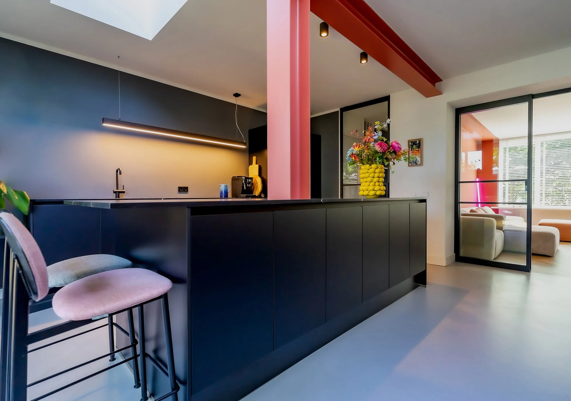 Donkerblauwe keuken met zwart eiland, roze velvet barkrukken en roze kolom met donkerrode balk. Lineaire hanglamp boven het werkblad. Rechts stalen glasdeur met zicht op terracotta woonkamer. Lichtstraat.