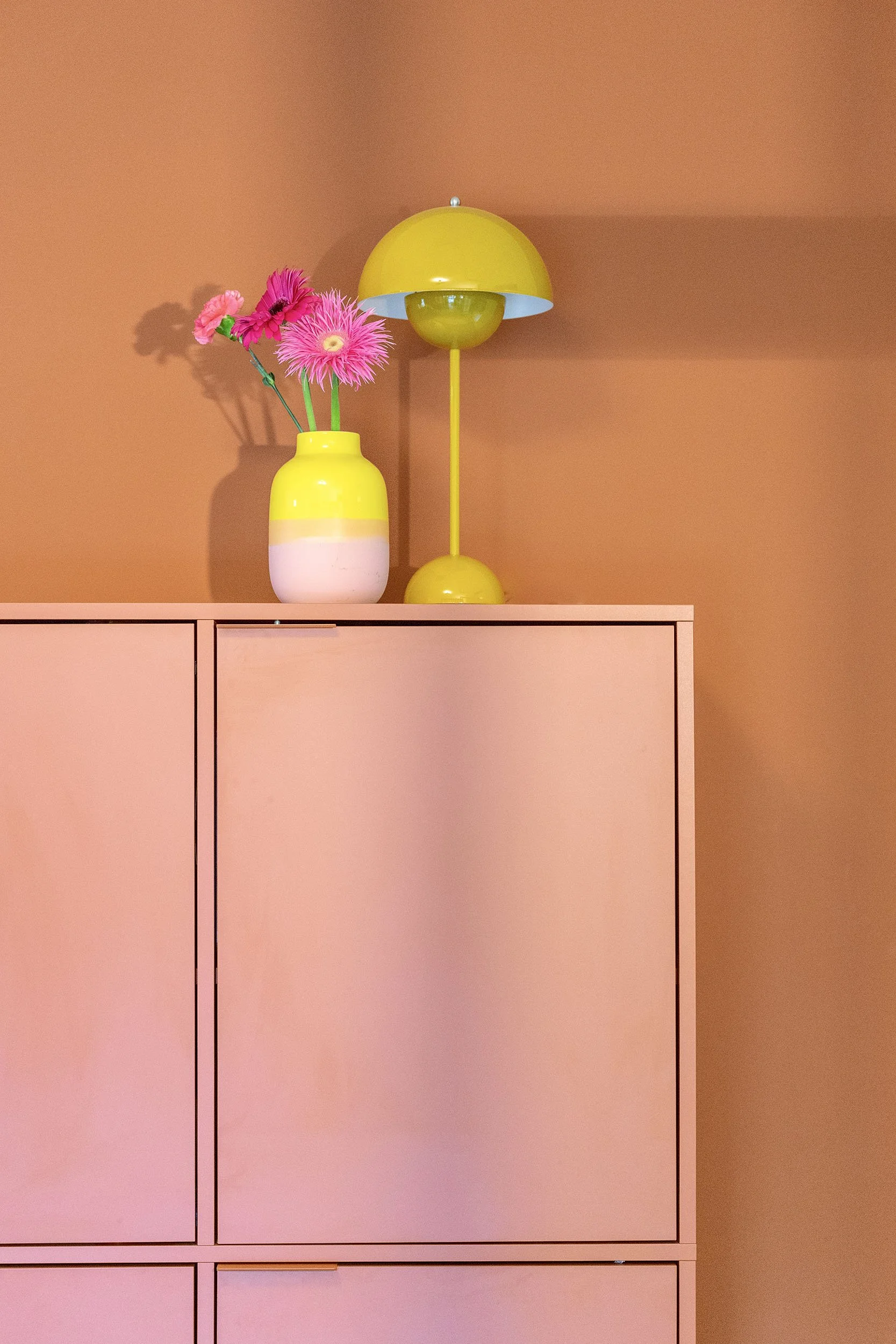 CharlotteMixt-interieurontwerp-Roze/blush modulaire kast met gouden greepjes, gele &Tradition Flowerpot tafellamp en tweekleurige geel-witte vaas met roze gerbera's. Terracotta oranje wand op achtergrond.