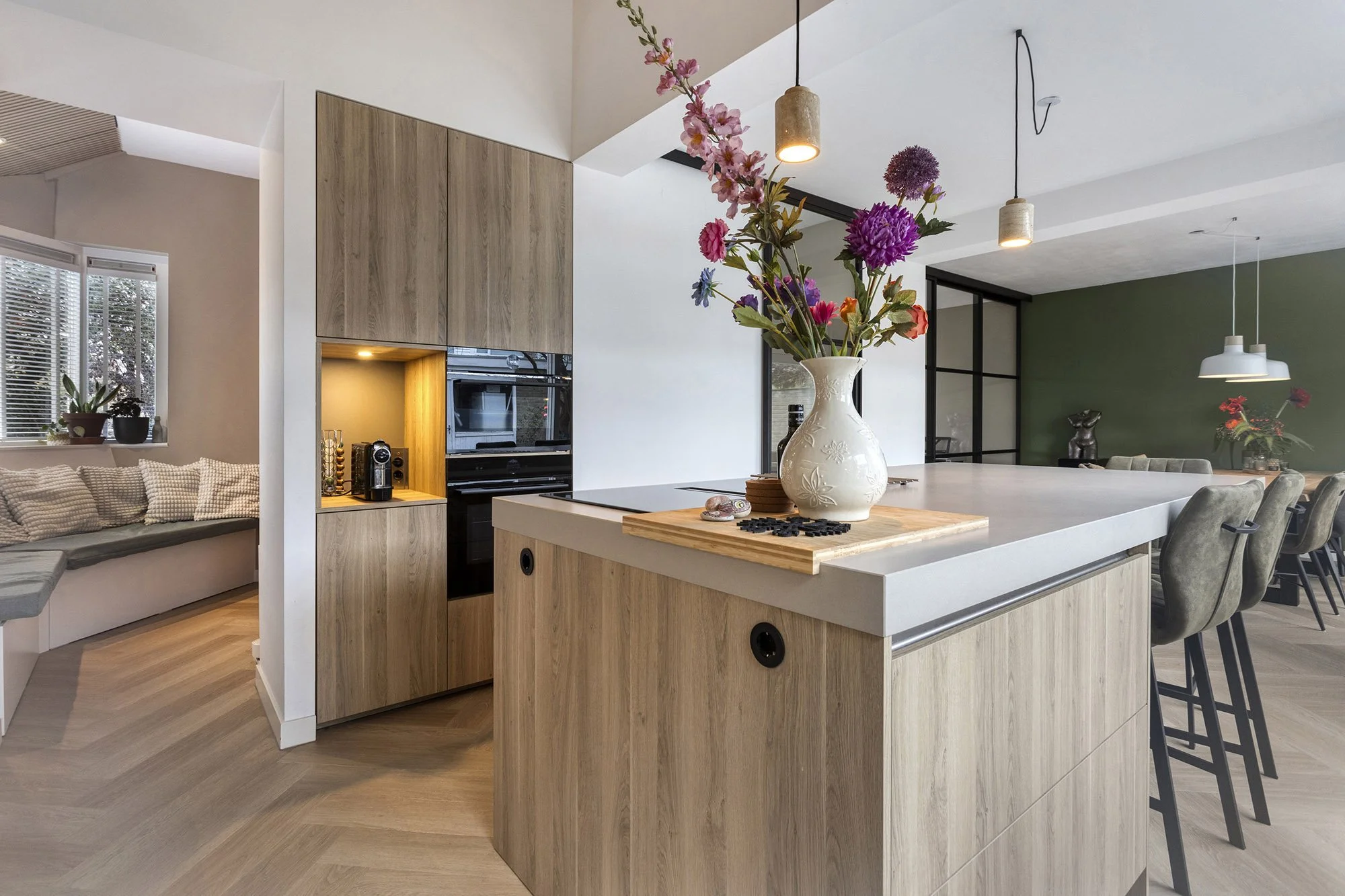 Eiken keuken met grijs werkblad, witte vaas met kleurrijk boeket en koffiehoek met zwarte inbouwovens. Links zithoek met bankstel. Op achtergrond donkergroene wand, stalen glasdeur en betonnen hanglampen boven eethoek met groene stoelen.