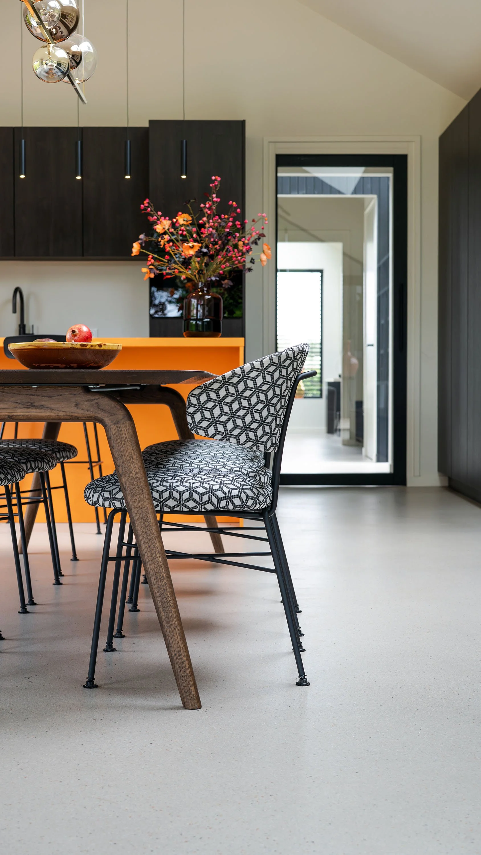 Zwart-wit geometrisch gestoffeerde eetkamerstoel naast houten tafel. Op achtergrond oranje keukeneiland met herfstboeket en donkere kastenwand. Deur met zicht op gang en bijgebouw.