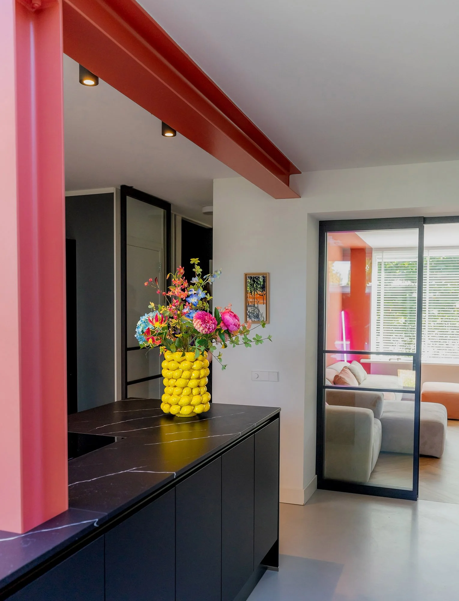 Woonkeuken Vught. Zwart marmeren keukenblad met gele bobbel-vaas en kleurrijk boeket. Roze kolom en donkerrode balk aan het plafond. Rechts stalen glasdeur met zicht op woonkamer met terracotta wand.