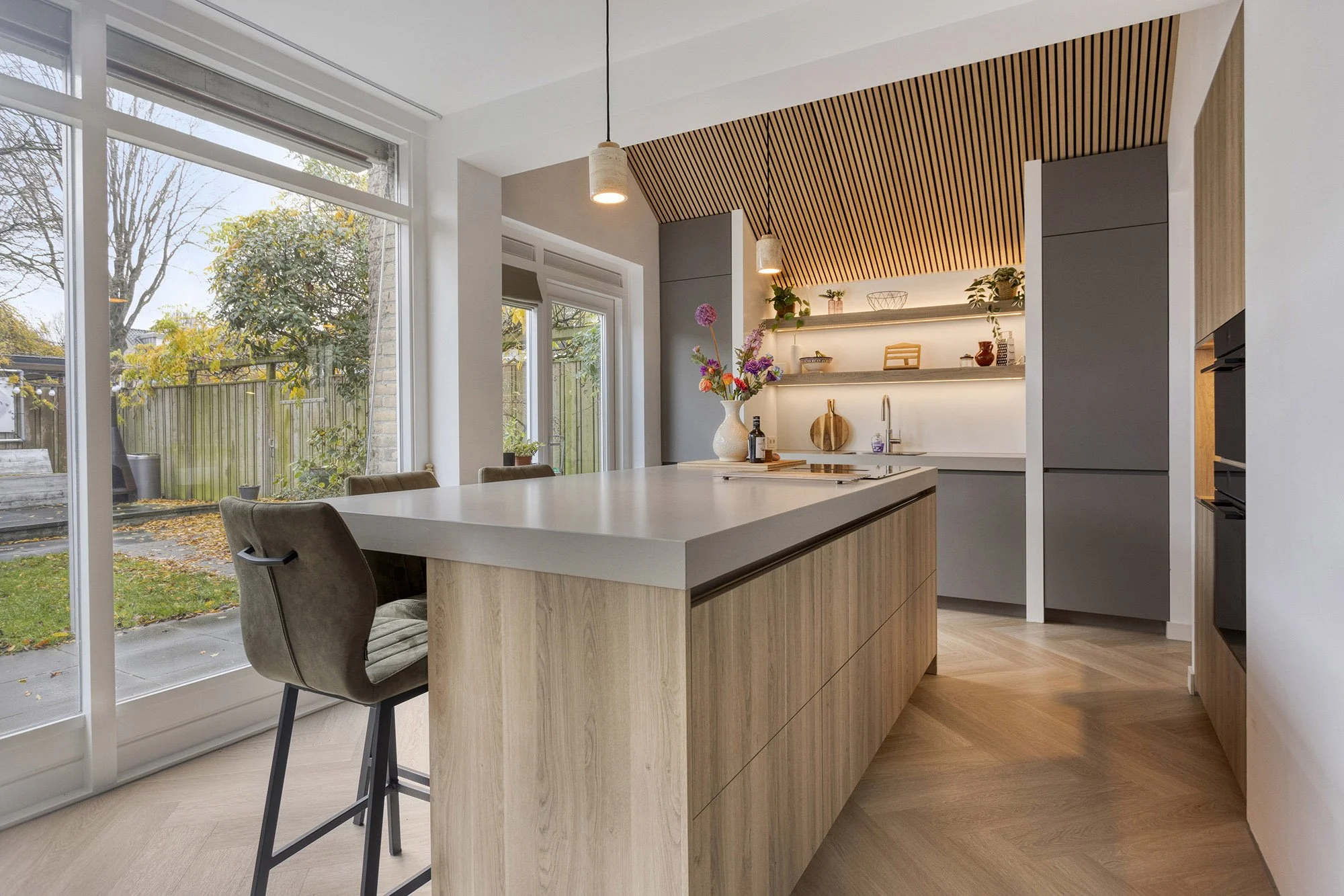 Lichte keuken met eiken eiland en groene velvet barstoel met zwart frame. Grote ramen en openslaande deuren met uitzicht op herfstuin. Op achtergrond grijze kasten, witte vaas met bloemen en verlichte houten schappen onder lamellenplafond.