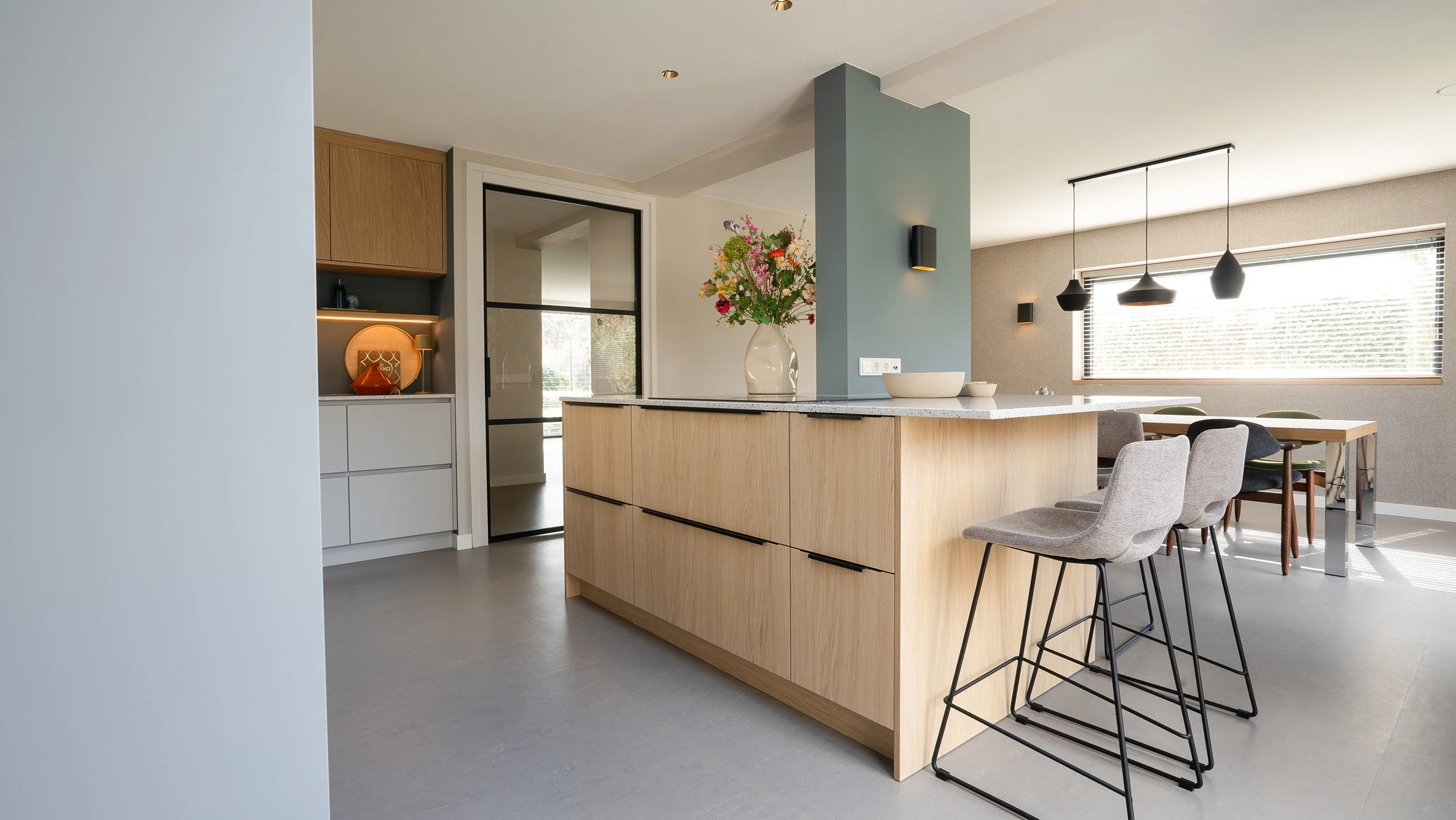 Moderne open keuken met een eiland van licht hout, bloemen in een vaas, barstoelen en een eetkamer met grote ramen en hanglampen.