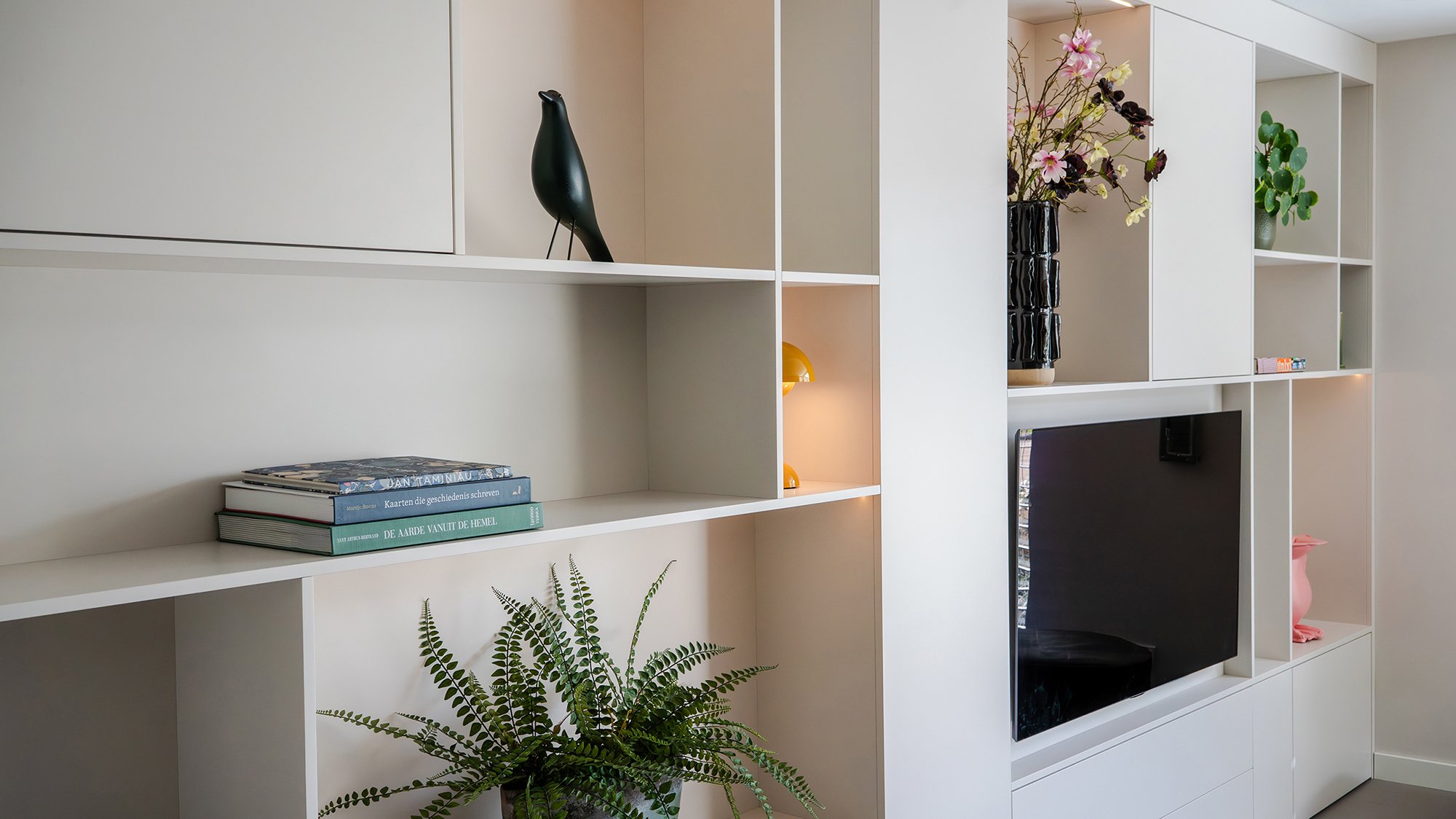 Detail witte wandkast met zwarte Eames House Bird, koffietafelboeken, varenplant, gouden tafellamp, donkere vaas met roze bloesem en ingebouwde televisie.