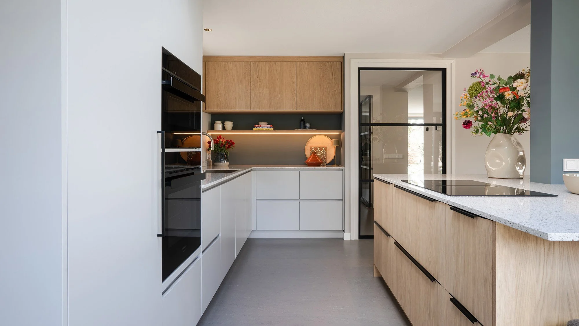 Interieurontwerp gezinswoning Vught. Keuken met Siemens ovens in witte hoge kast, grijs U-vormig keukenblok, eiken hangkasten en eiken eiland met inductiekookplaat en kleurrijk boeket.