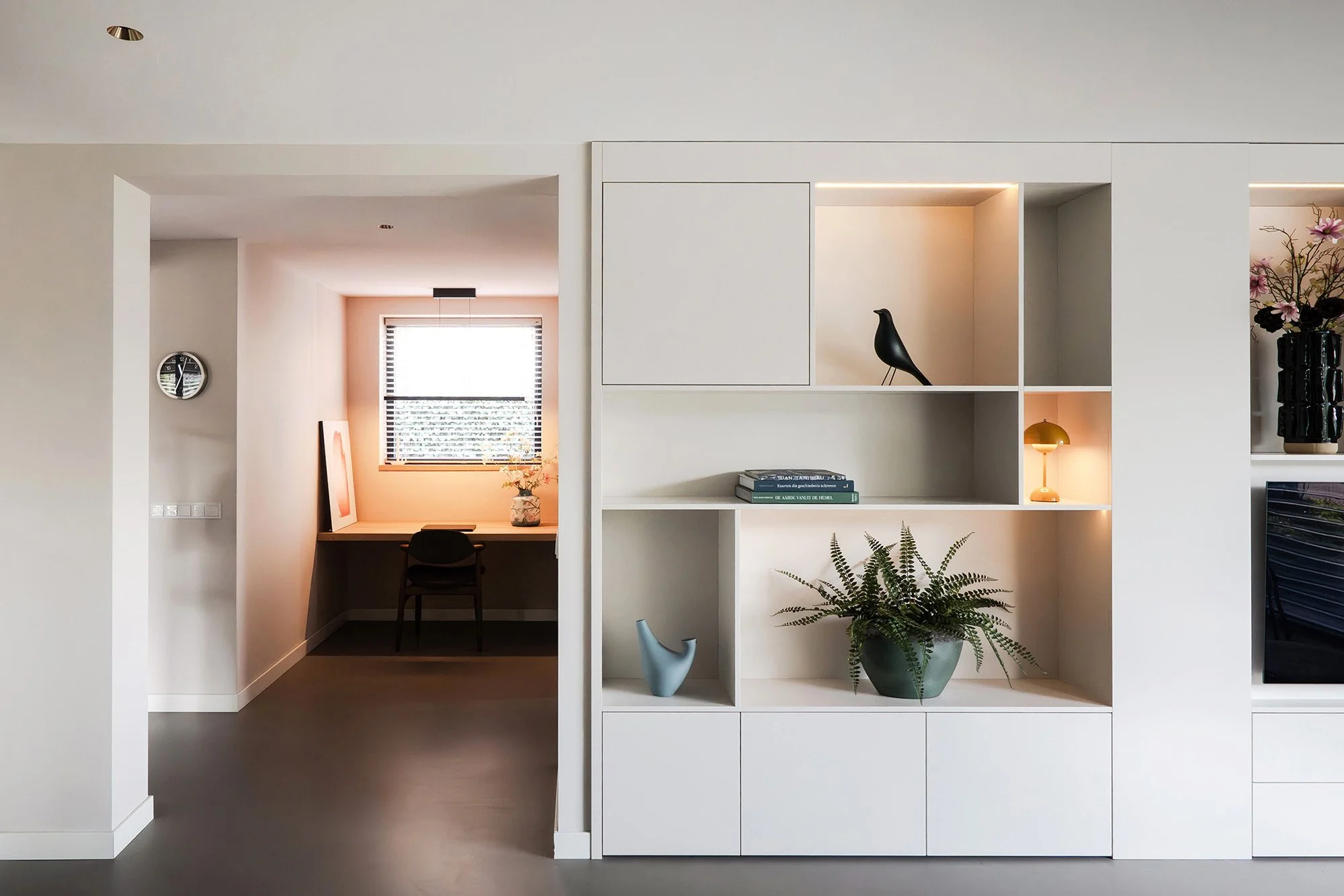 Woonkamer in Vught met witte wandkast met ledverlichting en decoraties. Links door opening werkniche zichtbaar met houten bureau, zwarte stoel en schilderij. Grijze betonvloer.