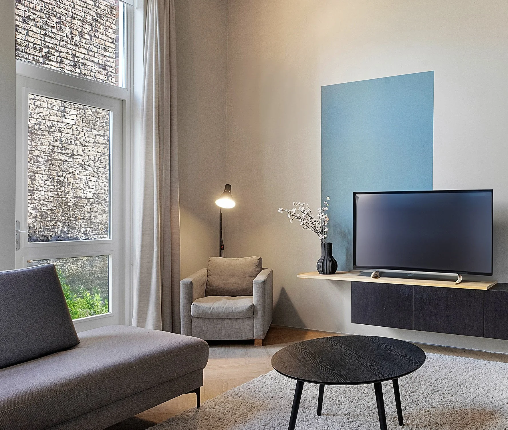 Woonkamer met donker zwevend tv-meubel, televisie en lichtblauw kleurvlak op wand. Grijze fauteuil met staande lamp in hoek bij hoog raam met uitzicht op bakstenen muur. Rond zwart salontafel op vloerkleed. interieurontwerp van CharlotteMixt.