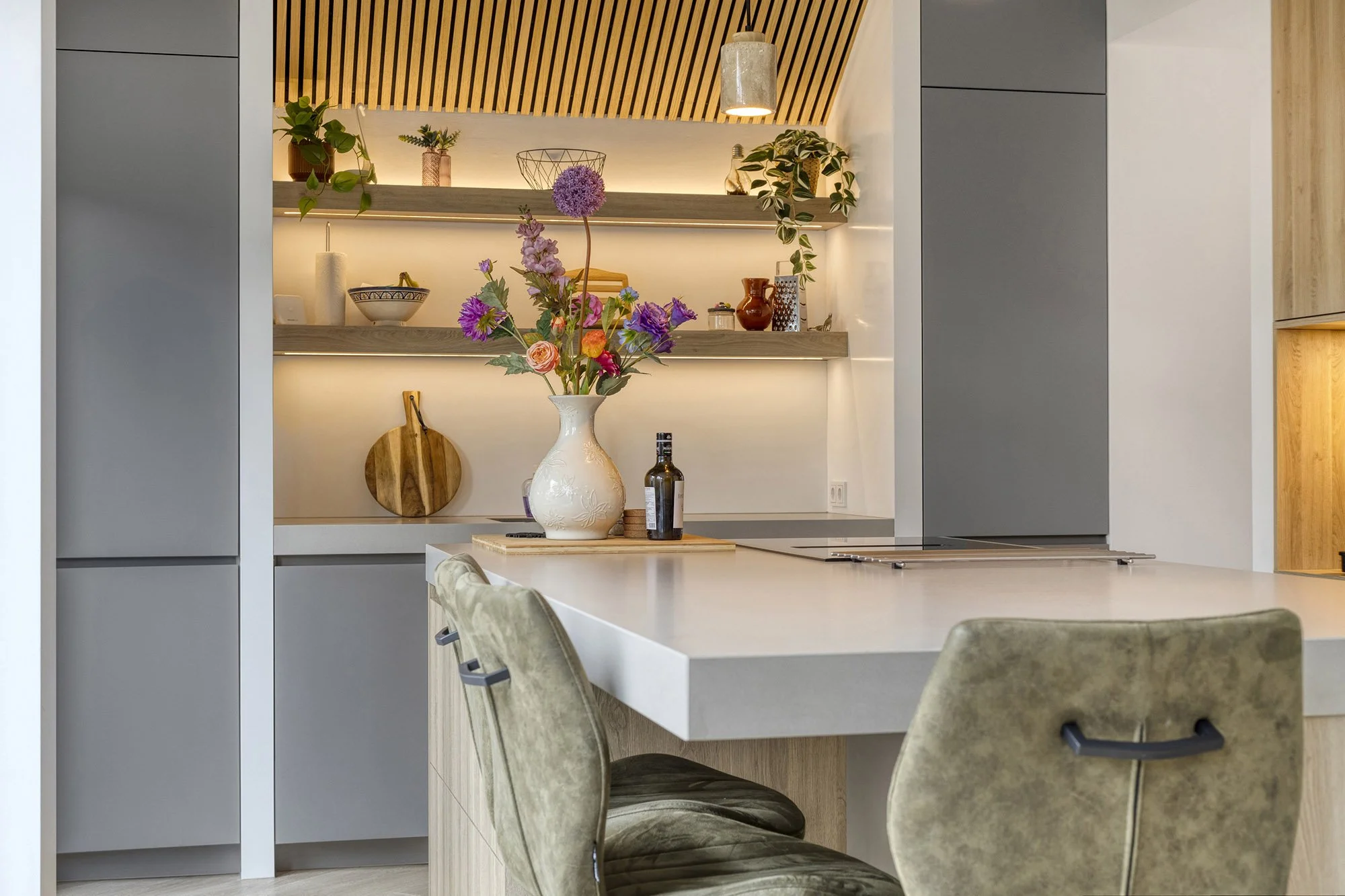 Zicht vanachter twee groene velvet barkrukken over het witte keukeneiland. Op het blad een witte vaas met kleurrijk boeket en olijfoliefles. Op achtergrond verlichte houten schappen met accessoires en lamellenplafond met betonnen hanglamp.
