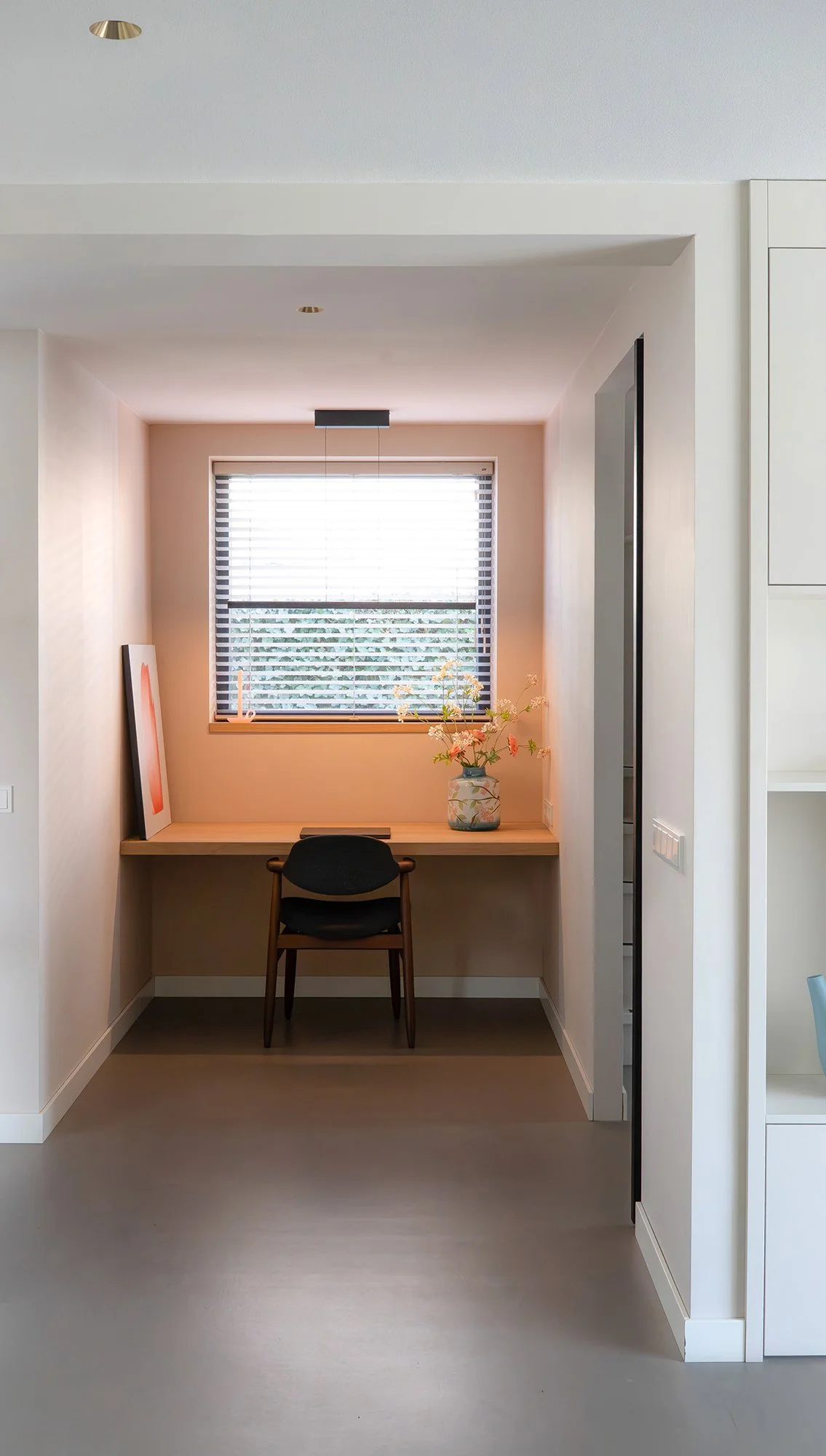 Interieurontwerp werkplek gezinswoning Vught. Ingebouwde werkniche met zalmroze wanden, zwevend houten bureau, raam met jaloeziën, zwarte houten stoel en vaas met roze bloemen. Rechts deel van witte wandkast en stalen deur zichtbaar.