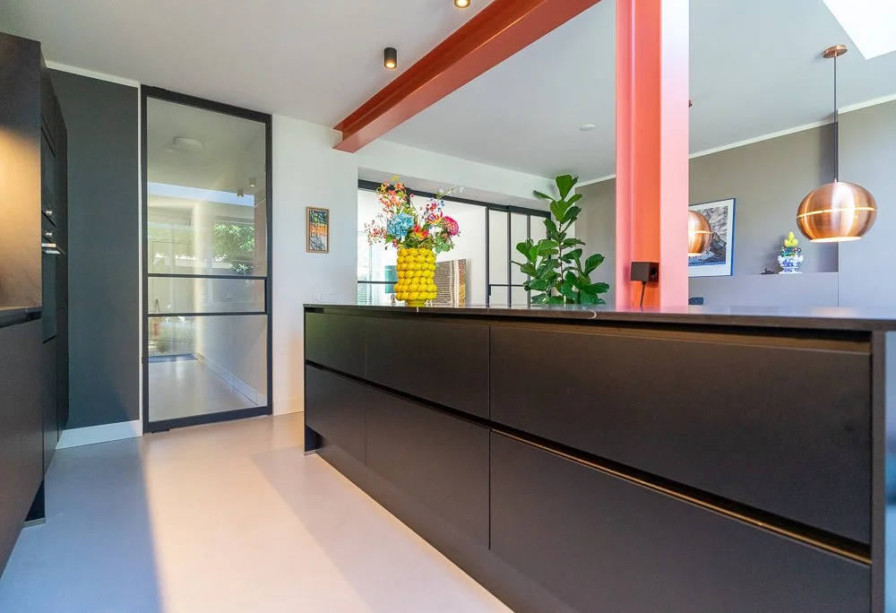 Zwart keukeneiland met gele bobbel-vaas en kleurrijk boeket. Roze kolom met donkerrode plafondbalken en spotjes. Stalen glasdeur links. Op achtergrond grote ficus en koperen hanglamp boven eethoek.