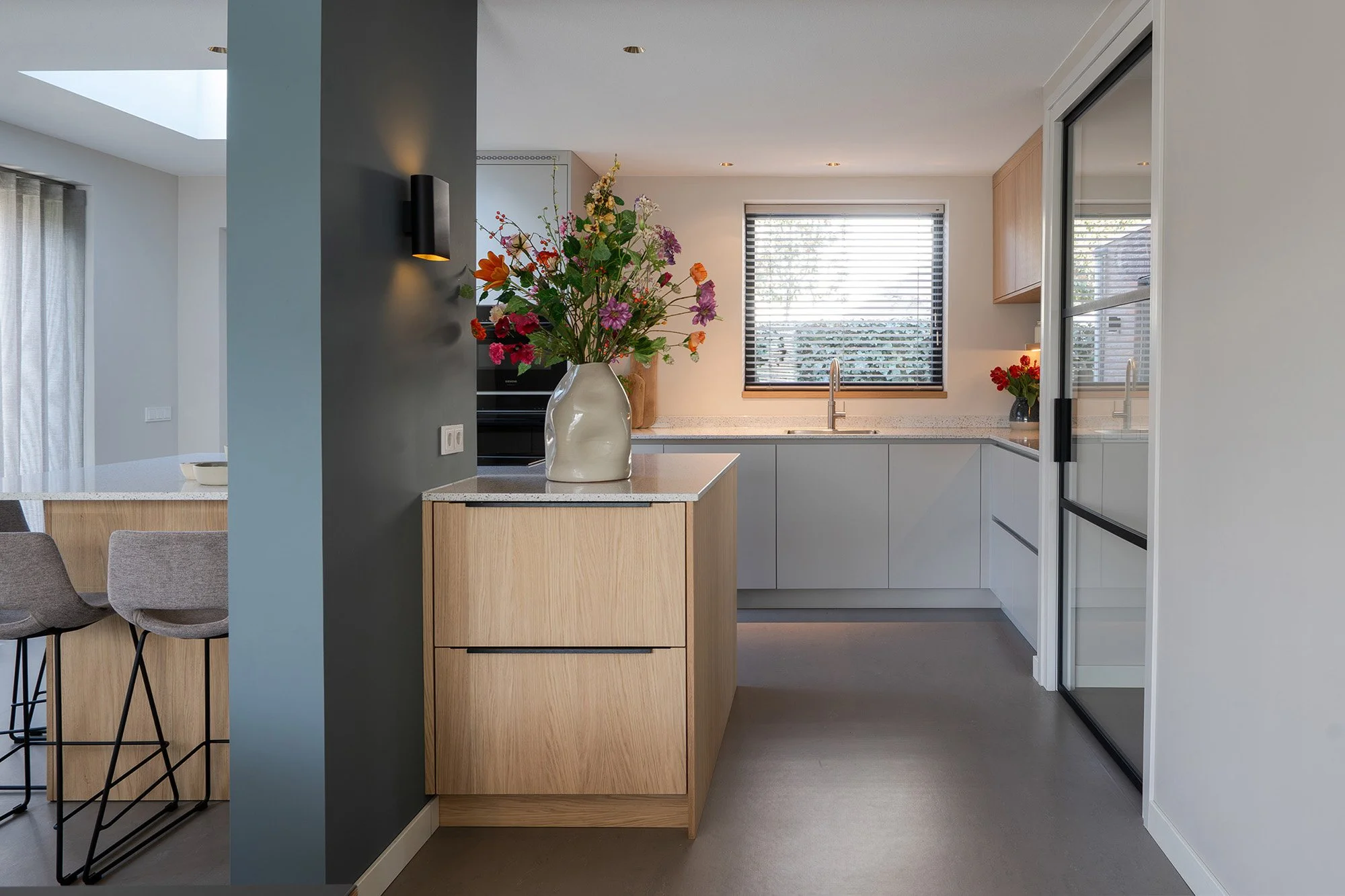 Interieurontwerp gezinswoning Vught. Keuken gezien vanuit woonkamer met blauwgrijze kolom, zwarte wandlamp en eiken eiland met witte vaas en kleurrijk boeket. Op achtergrond grijs keukenblok en stalen glasdeur.