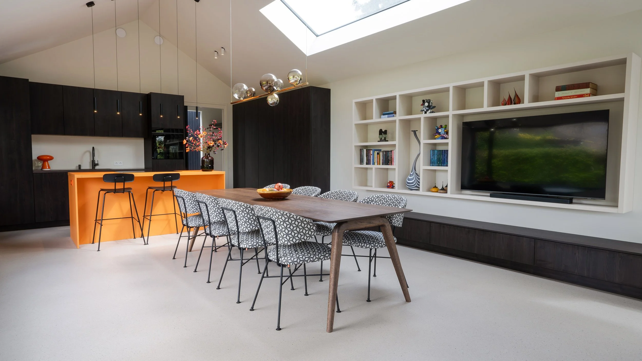 Eetkamer met lange houten tafel, zwart-wit geometrisch gestoffeerde stoelen en glazen bollampen. Rechts witte wandkast met tv en decoraties. Op achtergrond oranje keukeneiland en dakraam.