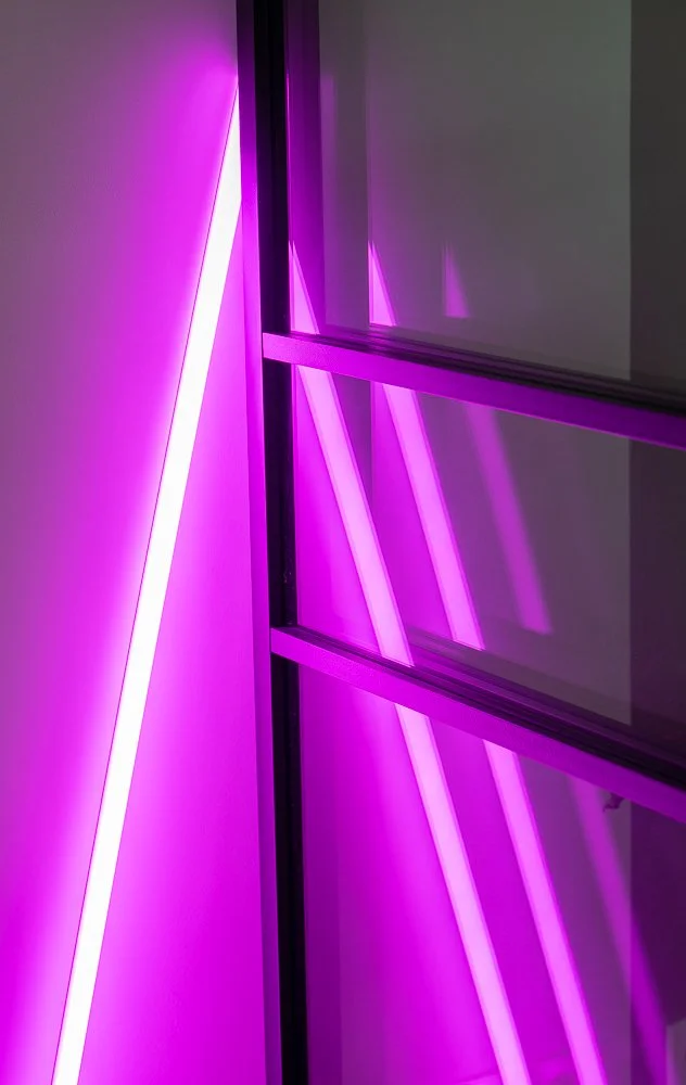 Abstracte close-up van een fuchsia roze neonlamp naast een stalen glasdeur. Het felle neontlicht werpt kleurrijke reflecties op de deur en de omliggende wand. CharlotteMixt