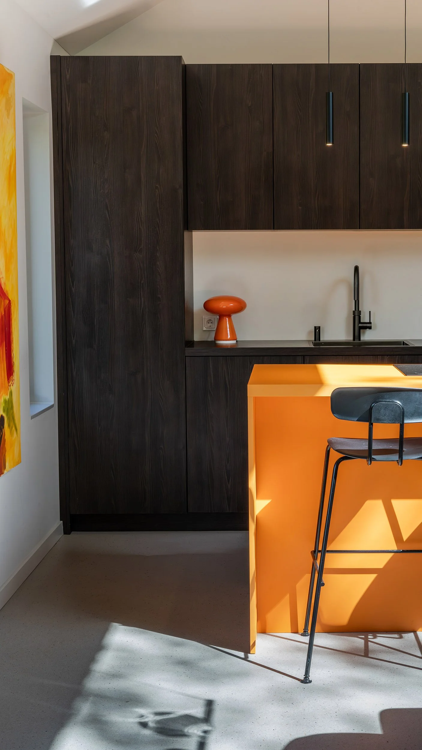 Donkere houten hoge kasten met zwart werkblad, zwarte kraan en oranje paddenstoellampje. Oranje keukeneiland met zwarte barstoel. Links figuratief kunstwerk. Grijze betonvloer.