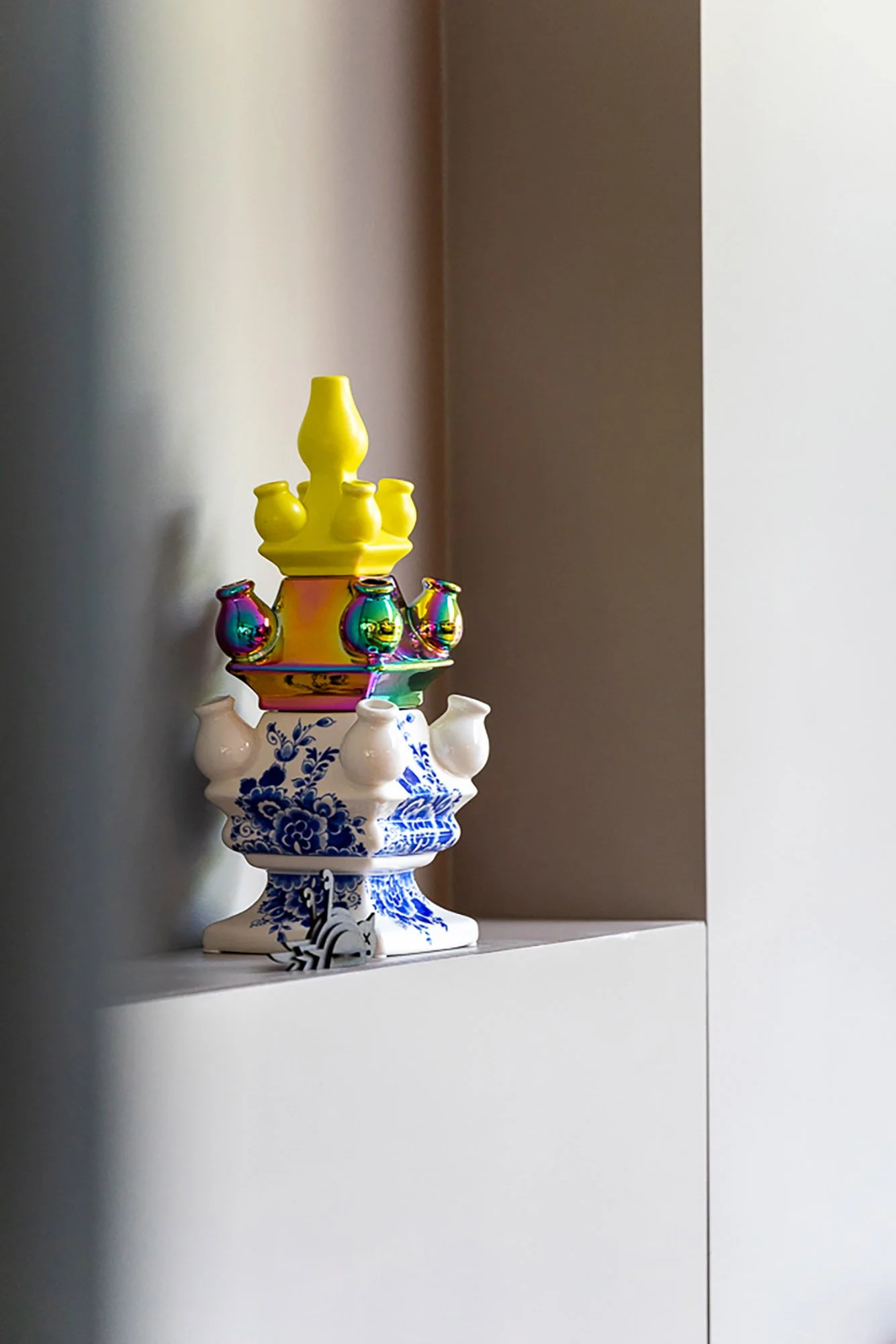 Decoratieve tulpenvaas in Delfts blauw en wit met een geel en iriserende toplaag, geplaatst in een taupe wandnis. Eigentijdse interpretatie van een klassiek Nederlands object.