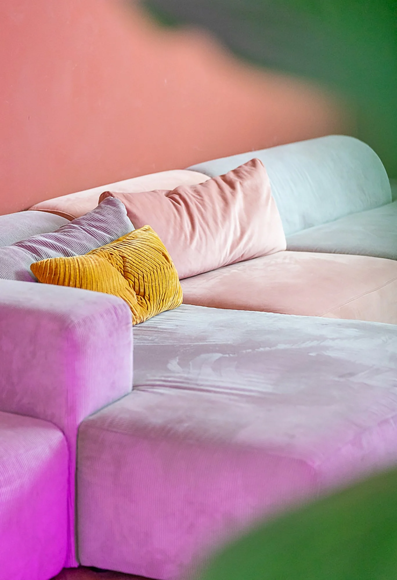 Detail van een grijs modulair bankstel met roze, geel en lila kussens voor een terracotta wand. Zachte pastelkleuren verlicht door magenta neonreflectie onderaan de bank.