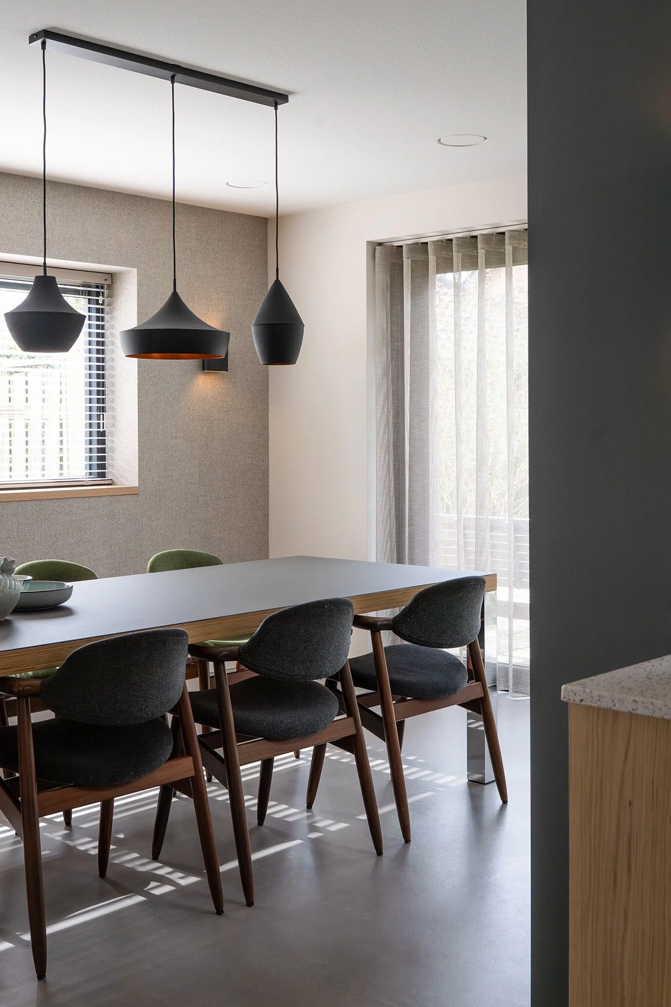 Interieurontwerp gezinswoning Vught. Eetkamer met donkergrijze eettafel, zes mid-century stoelen met houten poten en drie zwarte hanglampjes. Taupe textuurrwand, raam met jaloeziën en grote ramen met linnen gordijnen.