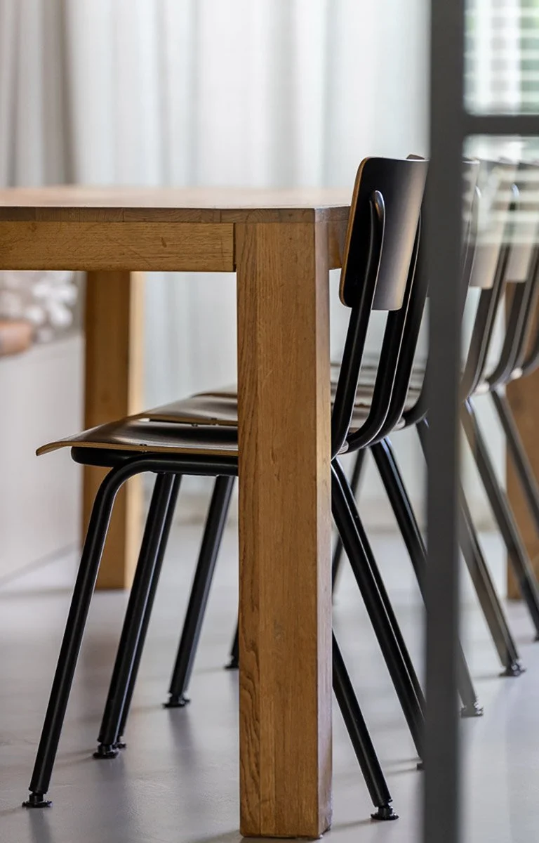 Close-up van zwarte stapelstoelen met metalen frame naast een houten eettafelpoot. Op de achtergrond witte vitrage en raam met jaloeziën. Grijze betonvloer. CharlotteMixt