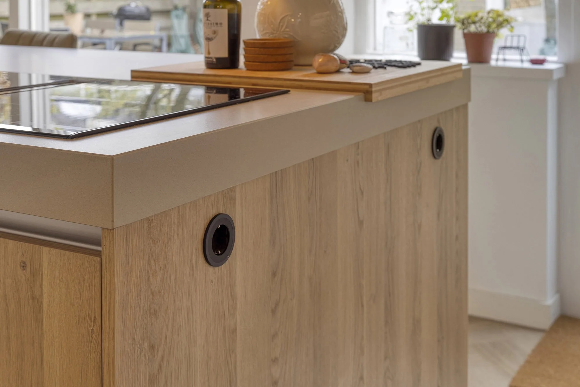 Close-up van de hoek van het eiken keukeneiland met grijs werkblad en zwarte inductiekookplaat. In het frontje twee ronde zwarte inbouwstopcontacten. Op het blad houten snijplank met accessoires. Op achtergrond raam met vensterbank en planten.