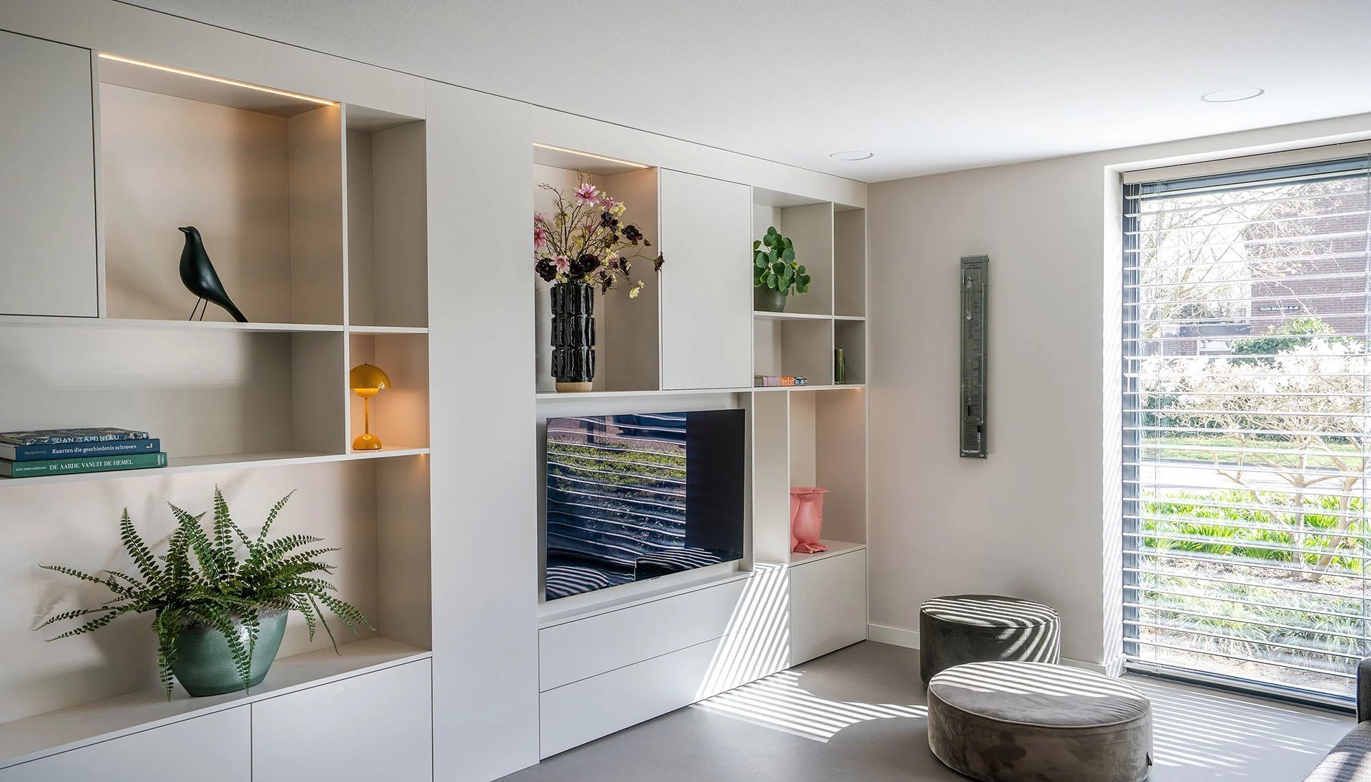 Woonkamer in Vught met grote witte maatwerk wandkast met open vakken, ingebouwde tv, Eames-vogel, varenplant en boeken. Groot raam met jaloeziën en tuinzicht. Twee ronde poefen op de vloer.