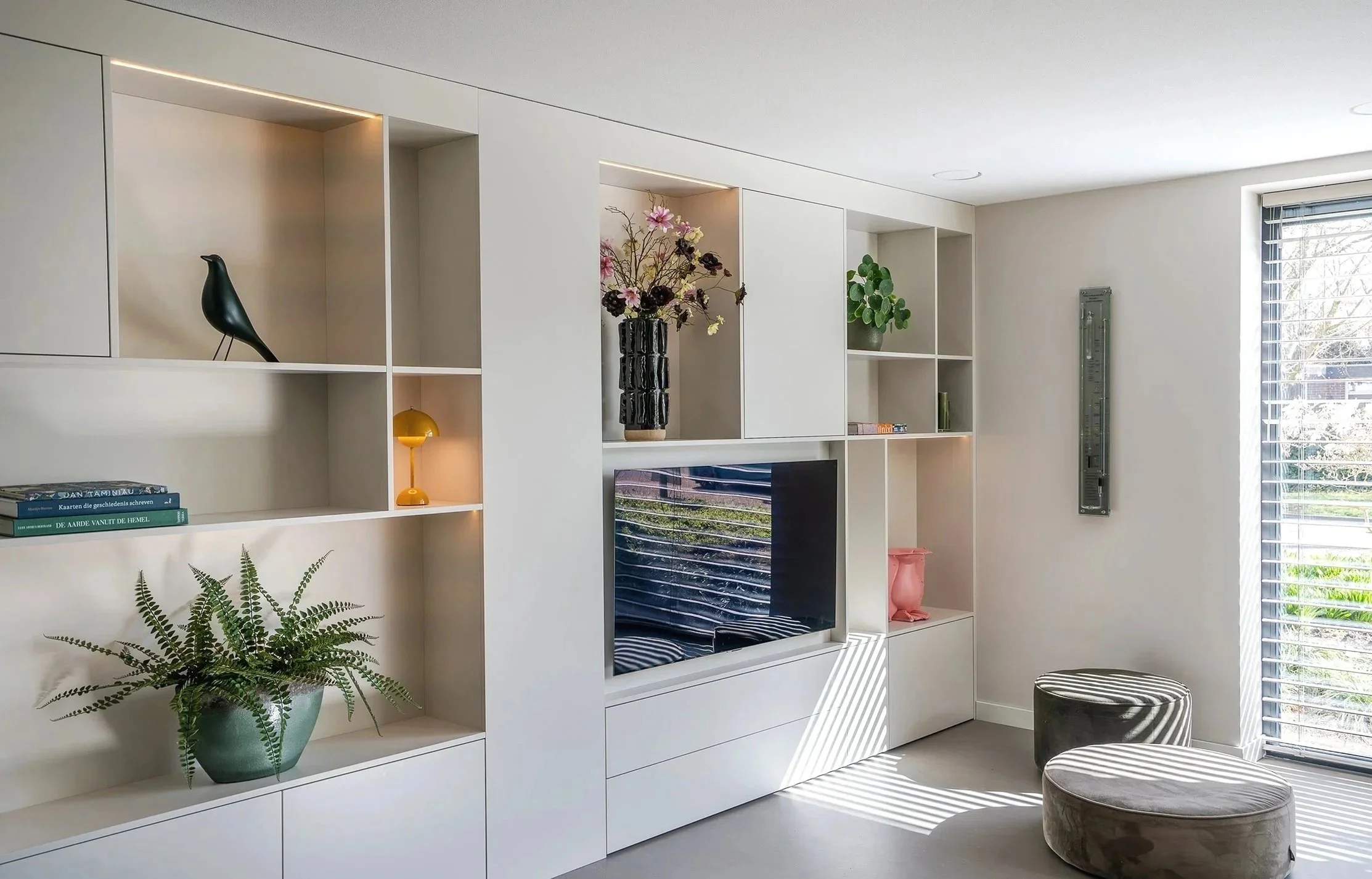 Moderne woonkamer met witte ingebouwde bibliotheek, planken met decoraties en planten, een tv, een raam met luiken die zonlicht toelaat, en twee zachte stoelen.