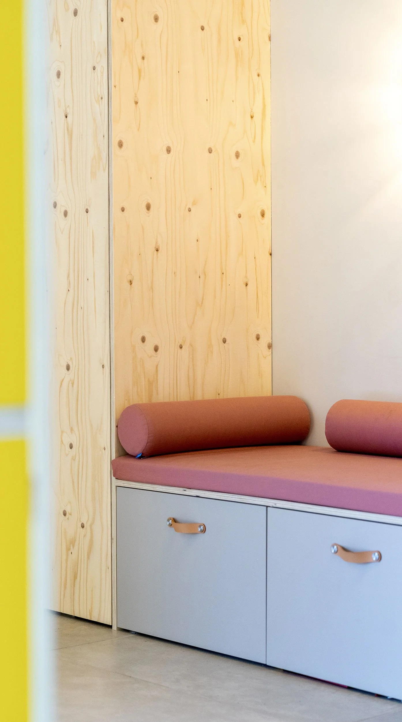 CharlotteMixt interieurontwerp Vught. Detail van roze gestoffeerde bankzitting met twee cilinderkussens, twee lade-opbergers met leren grepen, tegen een naturel multiplex wandpaneel. Links vleug geel zichtbaar.