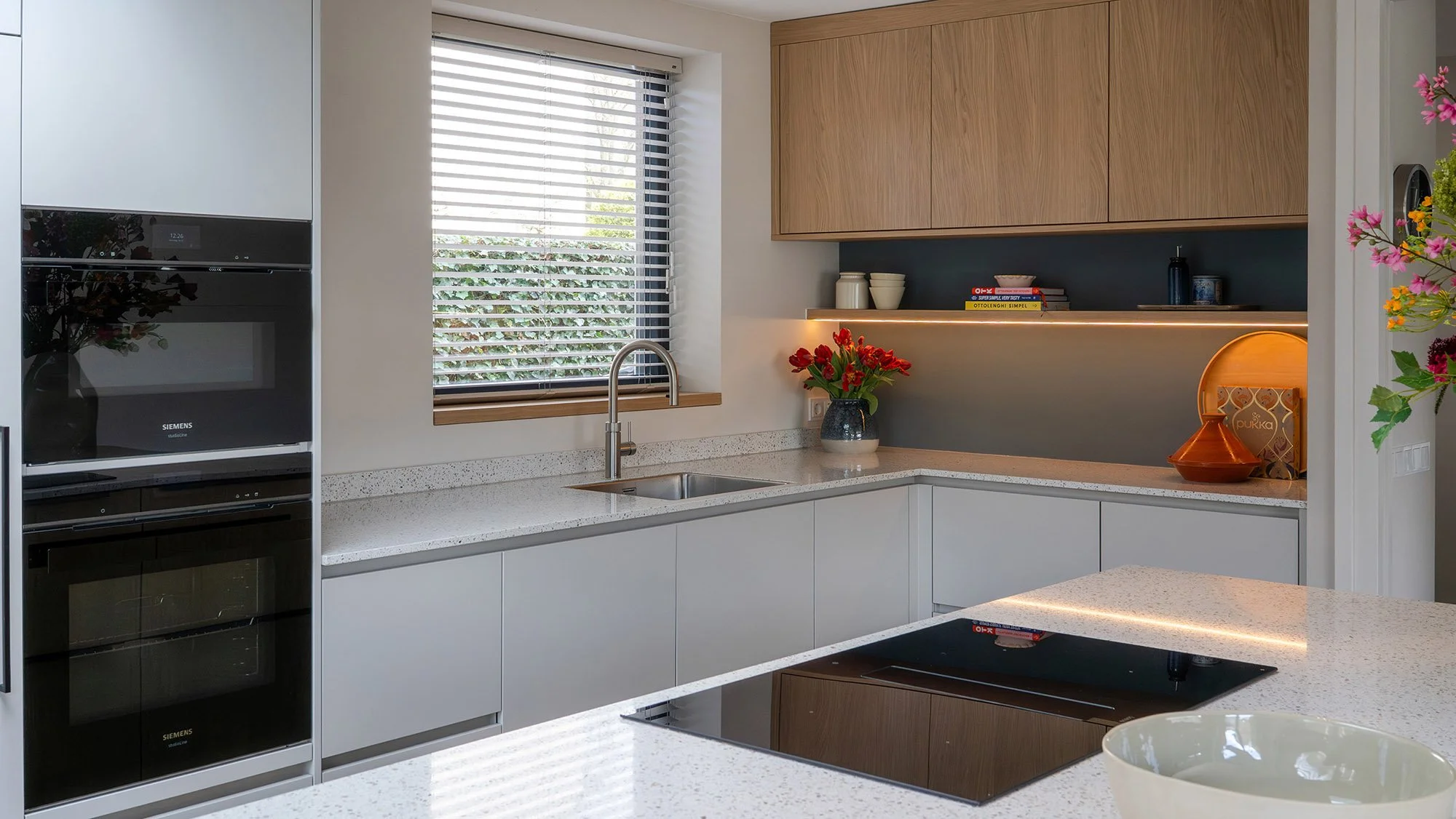 Interieurontwerp gezinswoning Vught. Keuken met twee Siemens ovens, grijs keukenblok met granieten werkblad, spoelbak, raam met jaloeziën en hoek met open schap, indirecte verlichting, rode tulpen en eiken hangkasten.