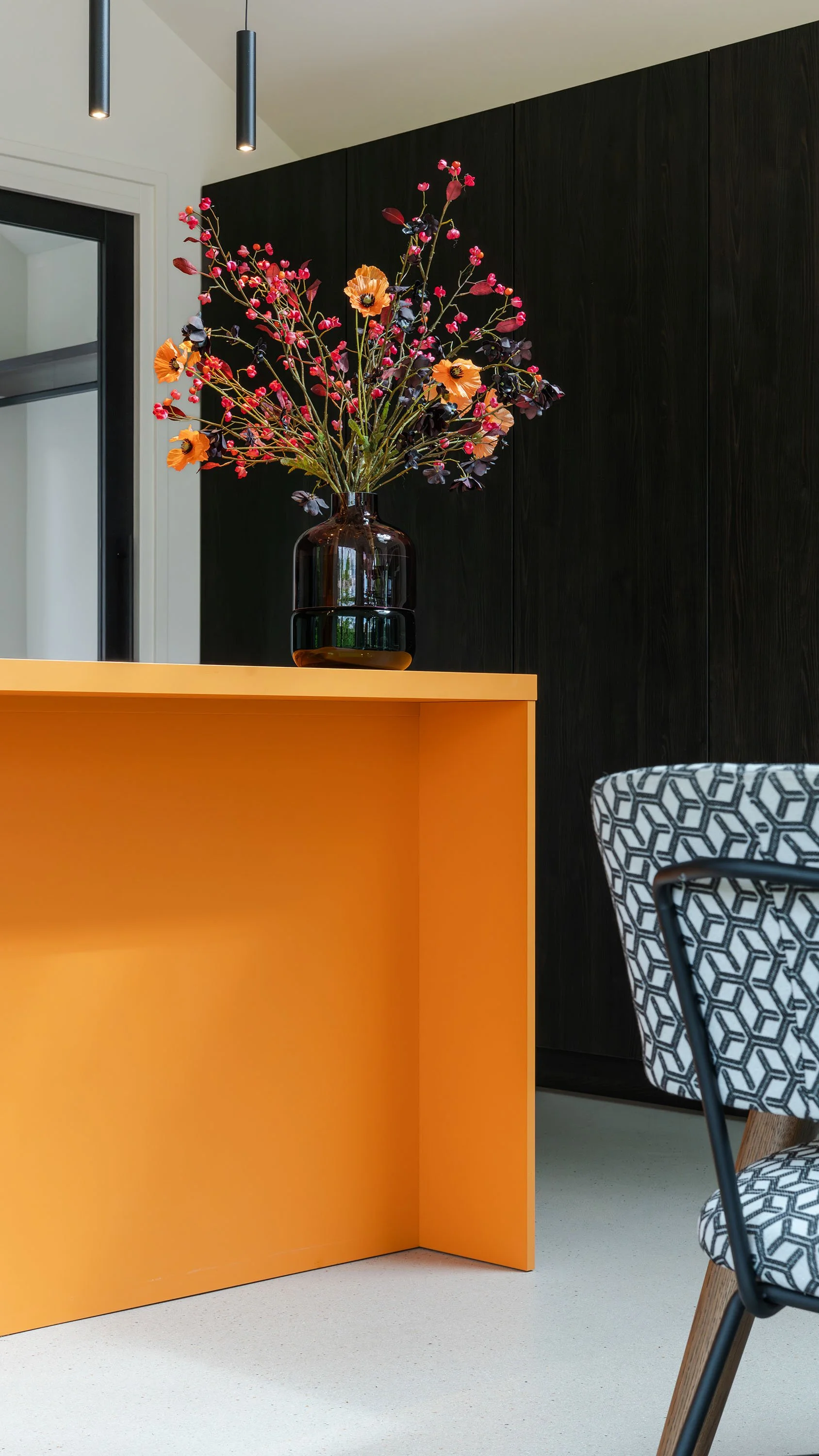 Een interieur met een gele tafel, een zwarte vaas met een grote bloemenarrangement, een zwart houten muur en een stoel met een geometrische zwart-wit patroon. Er hangen twee zwarte hanglampen aan het plafond.