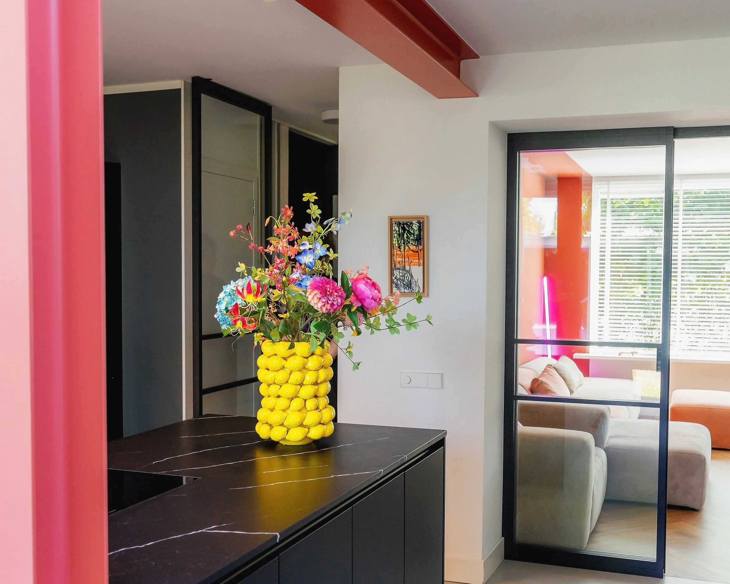Moderne keuken met zwart kookeiland en marmeren werkblad, opvallende rood-oranje stalen draagbalk, en een gele vaas met kleurrijke bloemen. Op de achtergrond een glazen schuifdeur naar een lichte woonkamer met zachte zitbank en neonverlichting.