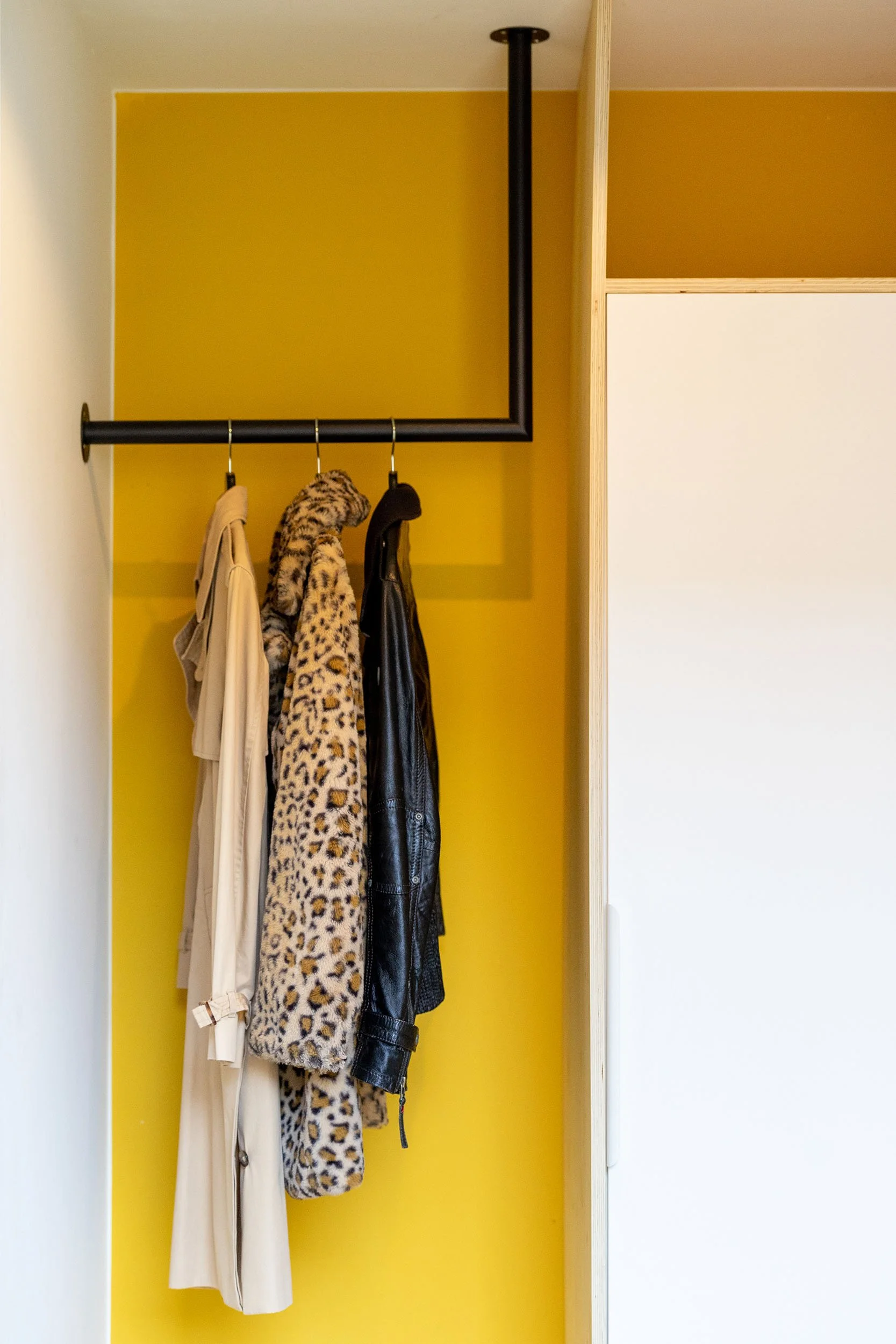 CharlotteMixt-interieurontwerp-Garderobe nis met mosterdgele achterwand en zwarte industriële plafondroede met drie hangende jassen. Rechts witte kastruimte en naturel multiplex afwerking.