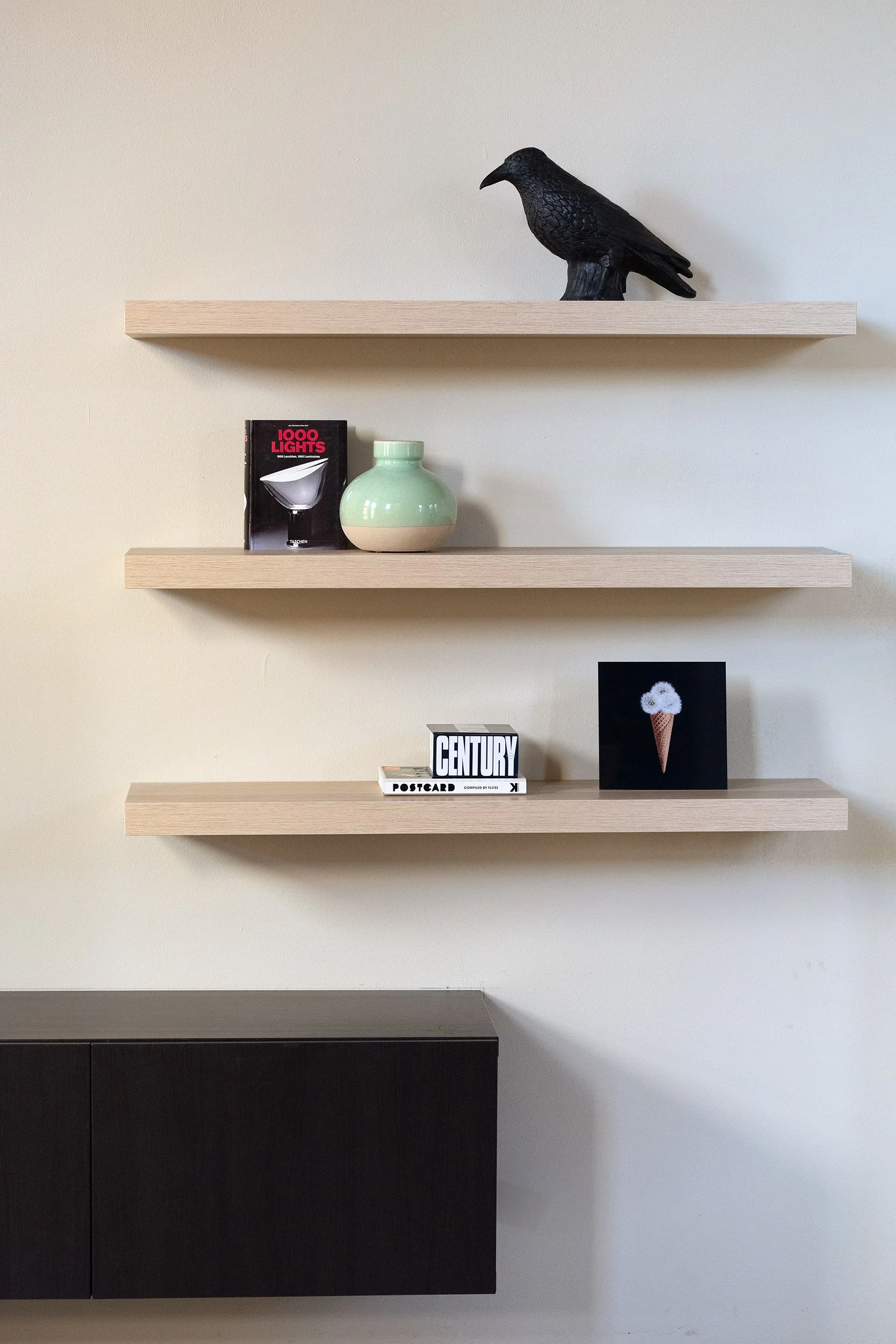 Drie zwevende eiken schappen op beige wand. Bovenste schap met zwarte kraai-sculptuur. Middelste met boek '1000 Lights' en mintgroene vaas. Onderste met boeken 'Century' en 'Postcard' en zwart kunstwerkje. Onder donker tv-meubel. CharlotteMixt