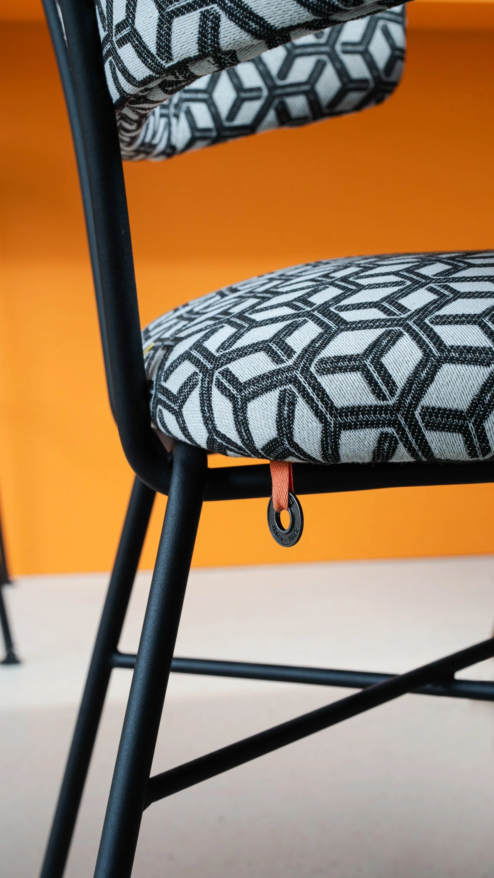 Close-up van zwart-wit geometrisch gestoffeerde eetkamerstoel met zwart metalen frame en oranje leren labellintje. Oranje keukeneiland als kleurrijke achtergrond.