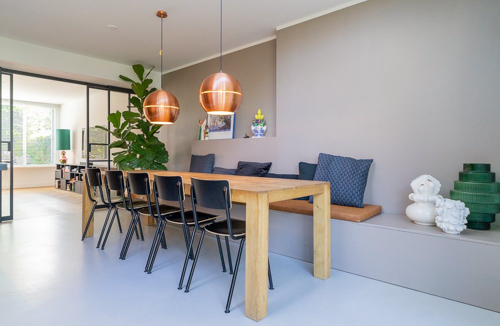Eetkamer met houten tafel, zwarte stapelstoelen en ingebouwde zitbank met blauwe kussens. Twee koperen bollampen, grote ficus en stalen glasdeur. Taupe wand met nis vol decoraties. Grijze betonvloer.