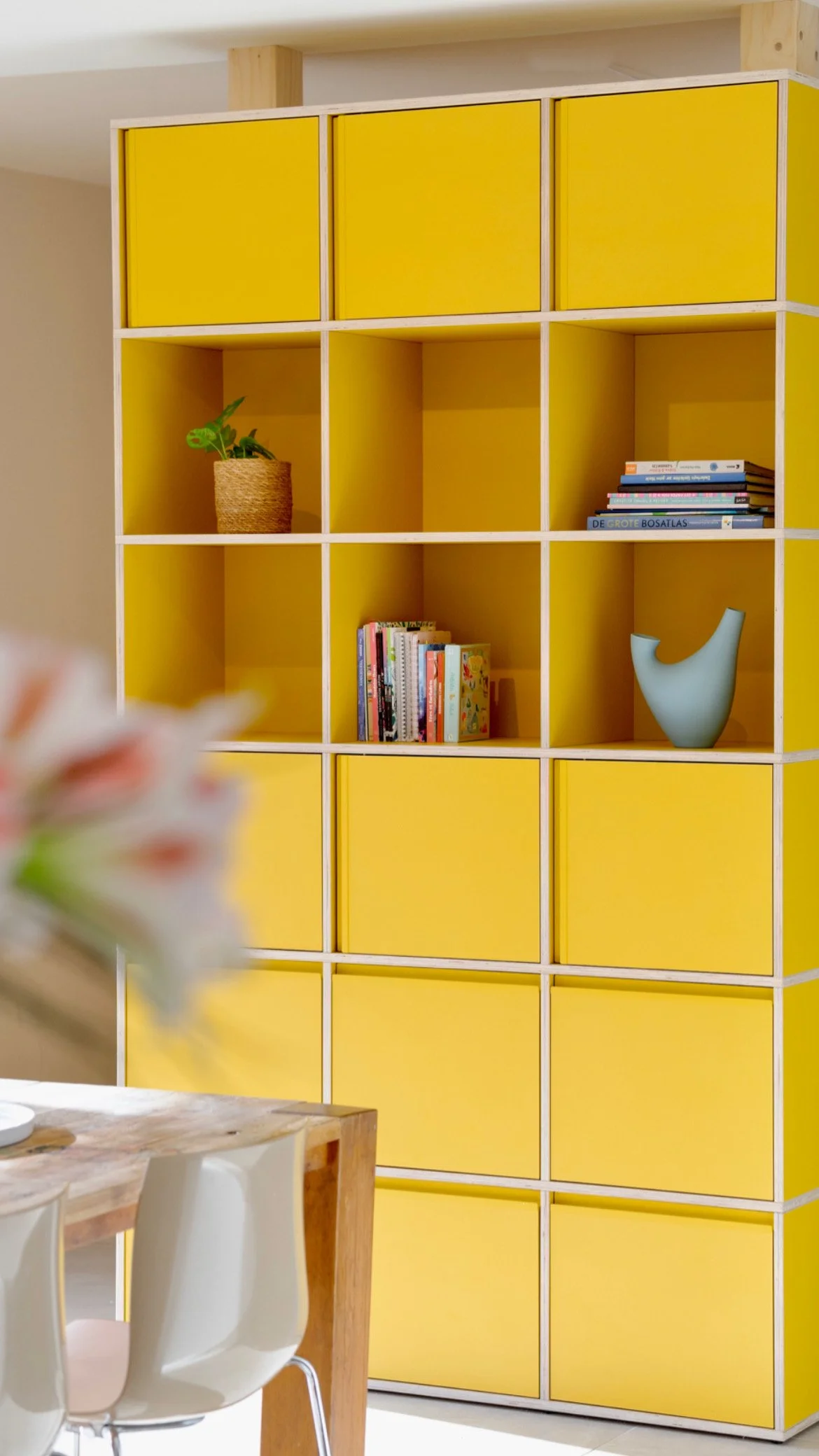 CharlotteMixt-interieurontwerp-Hoge gele modulaire kast als roomdivider, met open vakken met plant, boeken en blauwe vaas, en gesloten deuren. Multiplex kanten zichtbaar. Op voorgrond onscherpe bloemen en eethoek.
