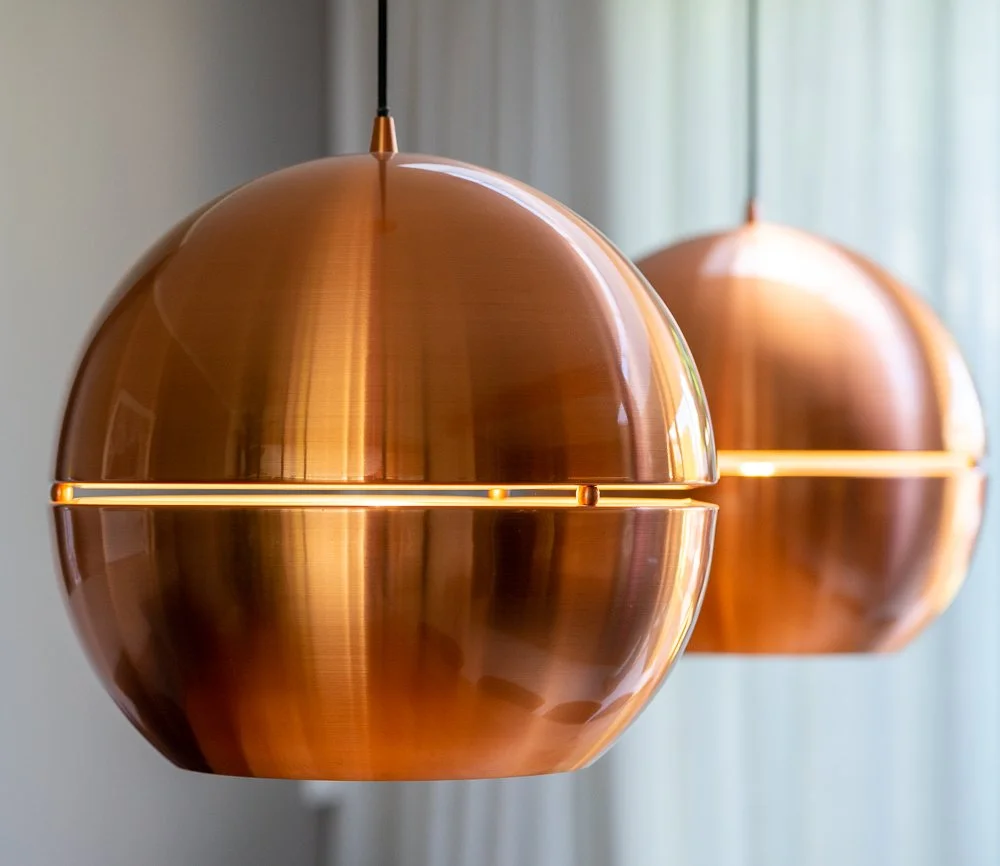 Close-up van twee grote koperen bollampen met een horizontale lichtgleuf in het midden. Warme gloed op het gepolijste koper. Onscherpe witte vitrage op de achtergrond. CharlotteMixt