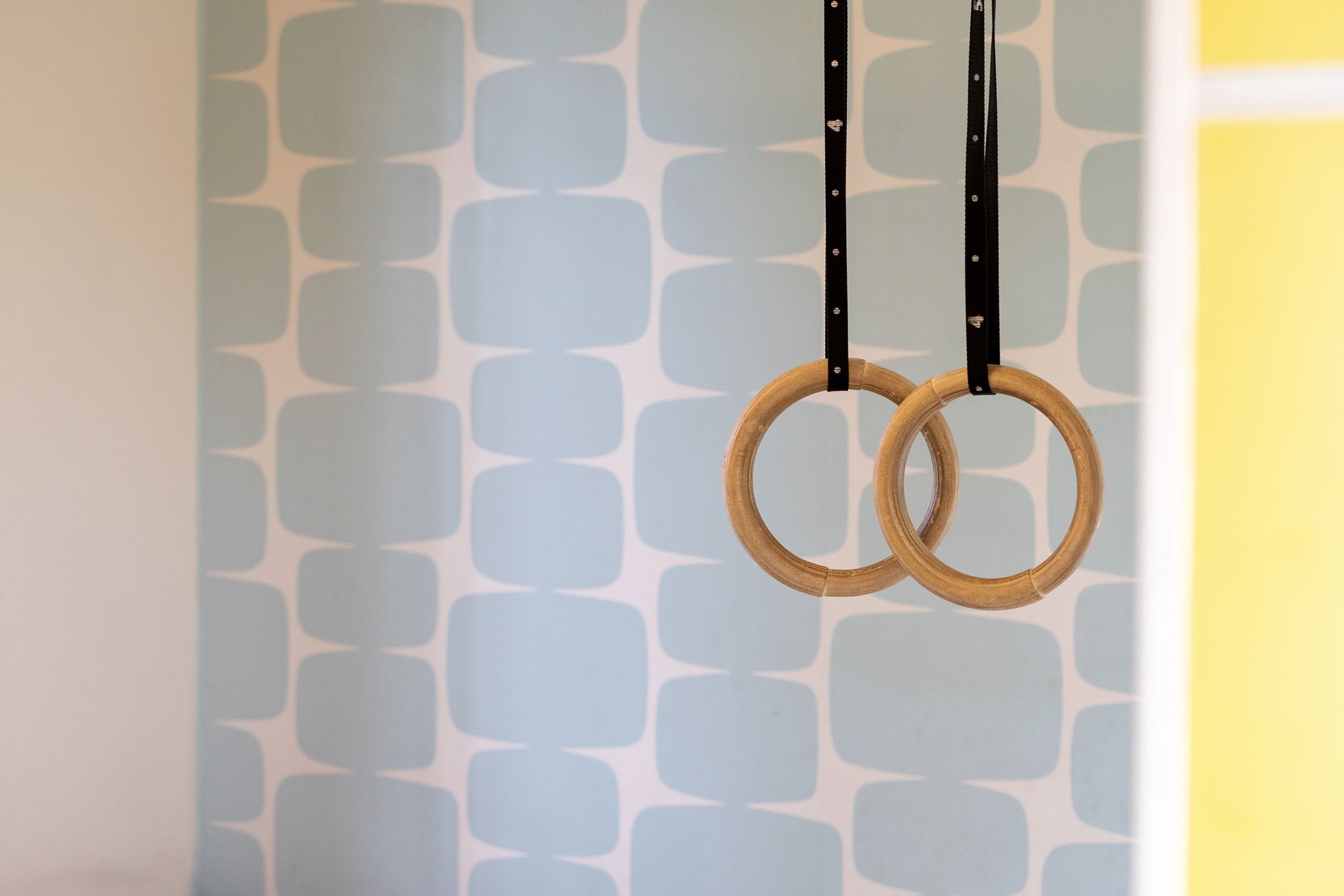 CharlotteMixt-interieurontwerp-Close-up van twee houten gymringen aan zwarte leren riemen, hangend voor lichtblauw geometrisch patroonbehang. Rechts een vleug geel van de boekenkast zichtbaar.