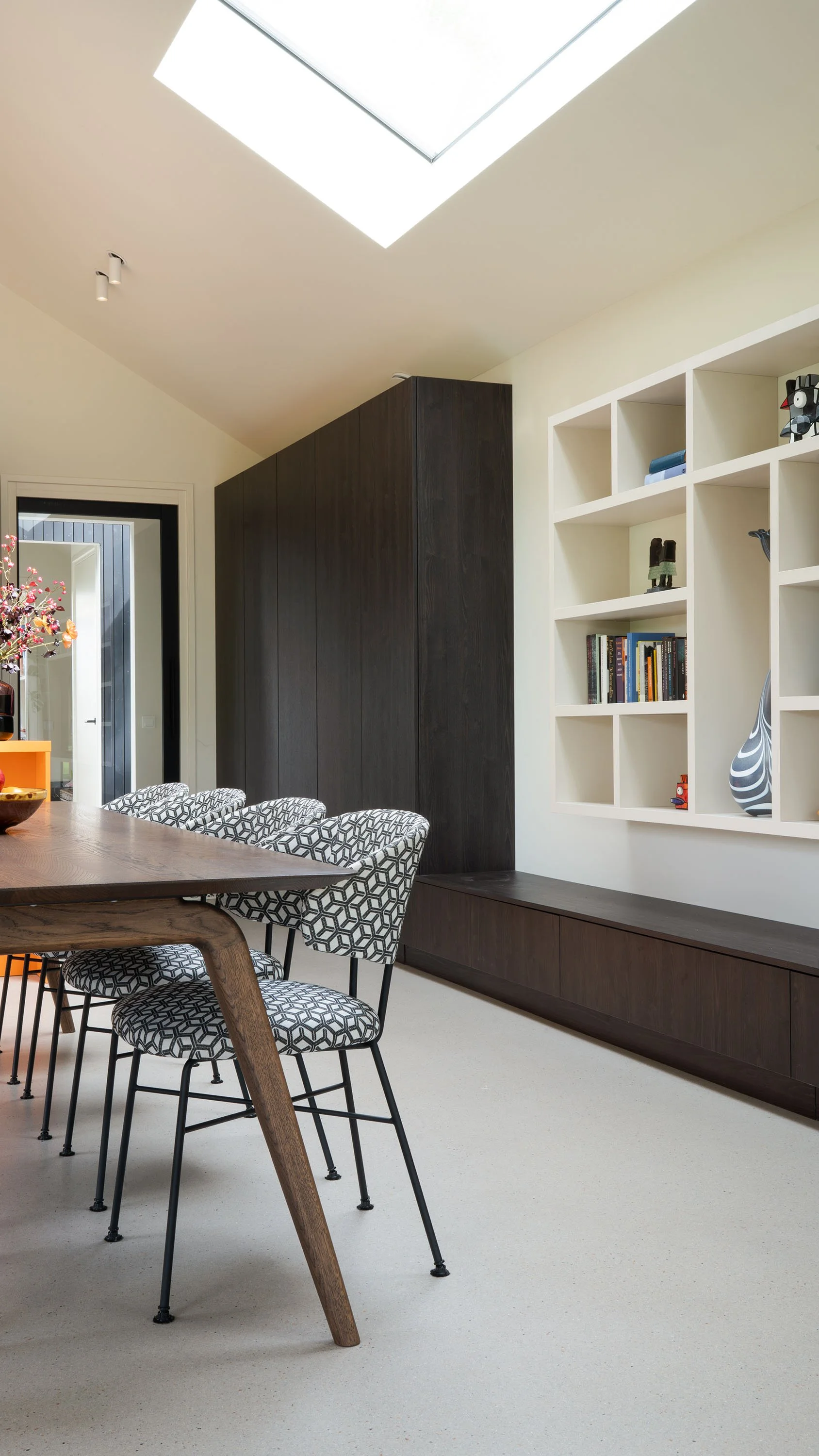 Zwart-wit geometrisch gestoffeerde eetkamerstoel naast houten tafel. Op achtergrond oranje keukeneiland met herfstboeket en donkere kastenwand. Deur met zicht op gang en bijgebouw.