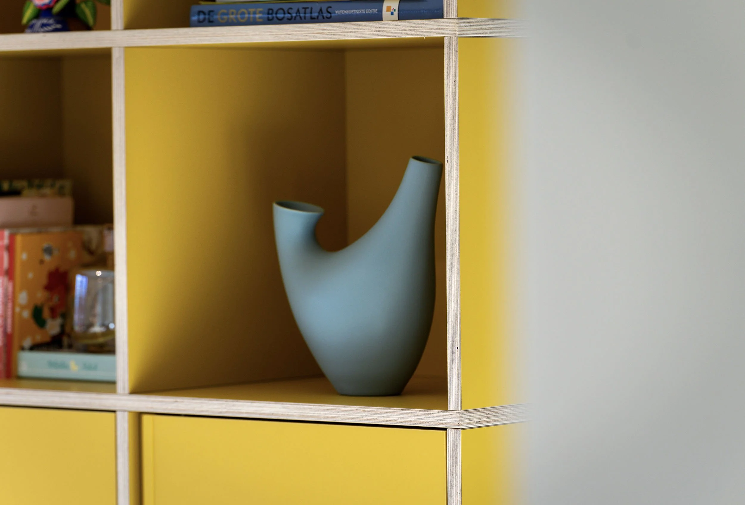 CharlotteMixt-interieurontwerp-Close-up van blauwe organische sculptuurvaas op plank van gele boekenkast met zichtbare multiplex kanten. Op achtergrond boeken en accessoires in aangrenzende vakken.