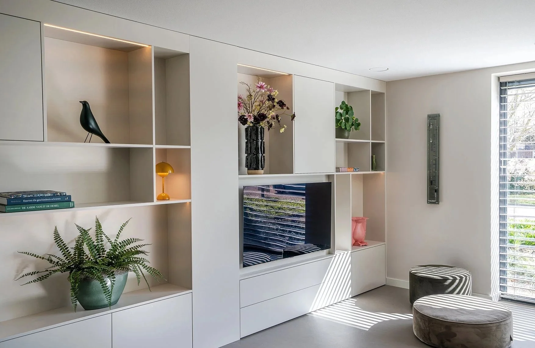 Woonkamer met witte open Planken kast, planten, bloemen, TV en zittsplek met een rond gestoffeerde poef en een stoel naast een raam.