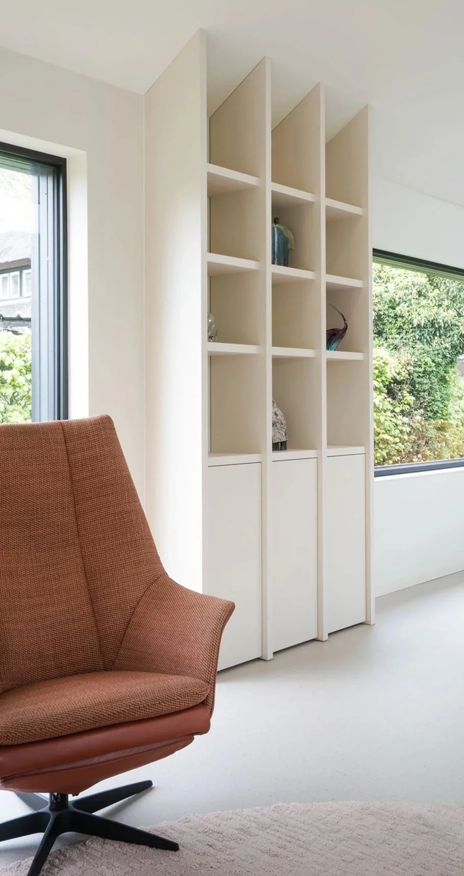 Detail van witte hoge maatwerk boekenkast met open vakken en blauwe keramische vaas. Oranje draaifauteuil op de voorgrond. Grote ramen met weelderig groen tuinzicht aan beide zijden.