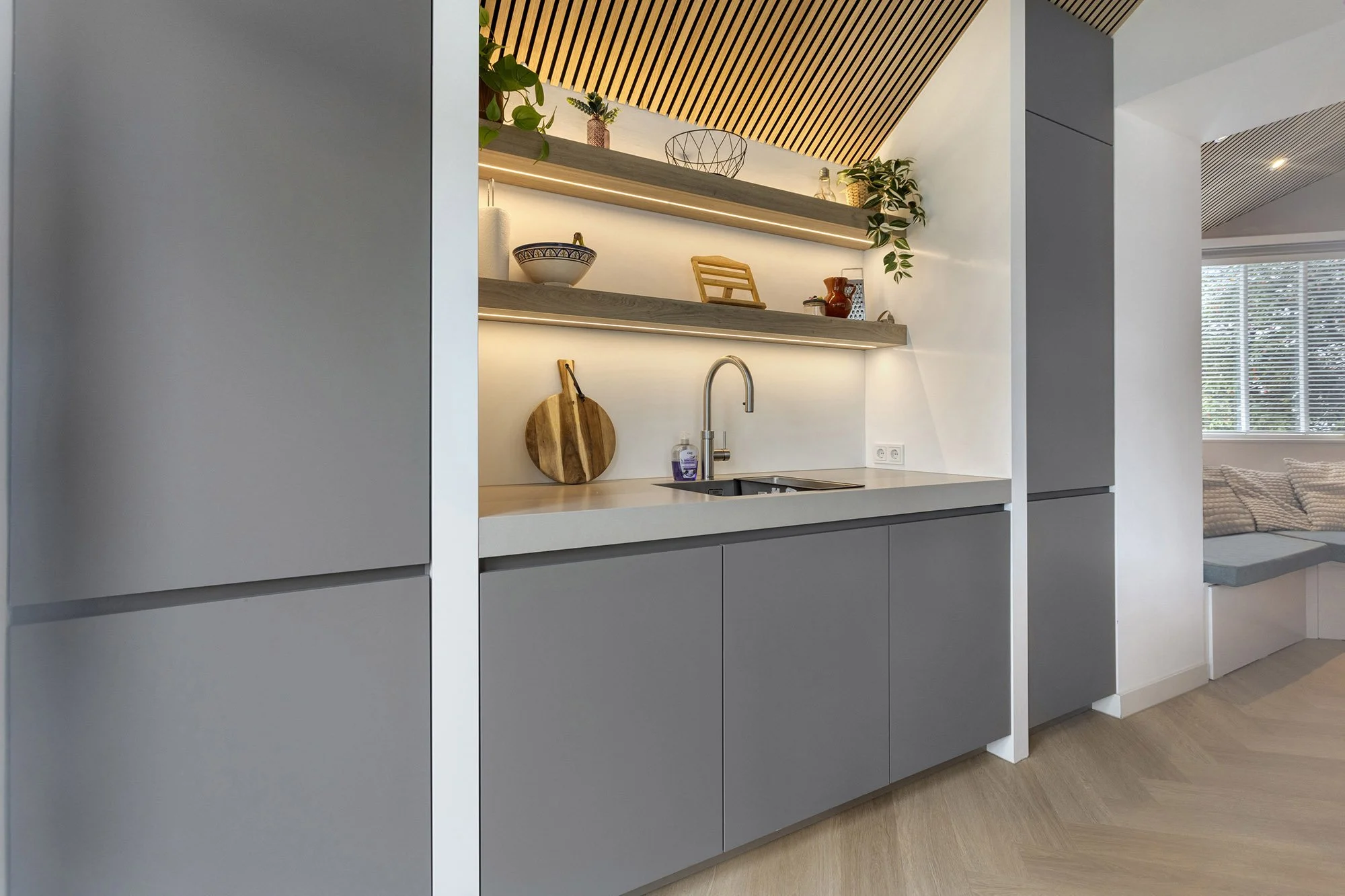 CharlotteMixt interieurontwerp Vught. Grijze keukenkasten met spoelbak, boogkraan en twee houten schappen met ledverlichting. Schappen gedecoreerd met planten, kommen, snijplanken en keukenaccessoires. Visgraatparket.