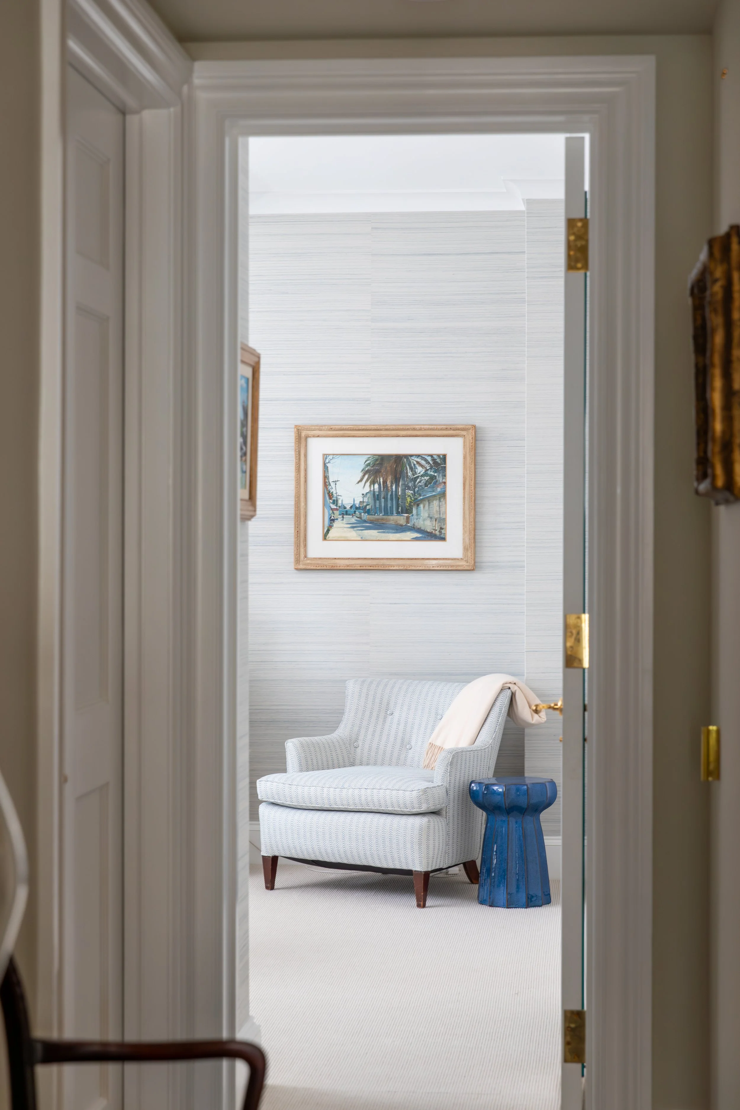 A cozy sitting area with a white upholstered armchair, a blue side table, and a framed painting of a street scene with palm trees hanging on a textured wall, seen through an open doorway.