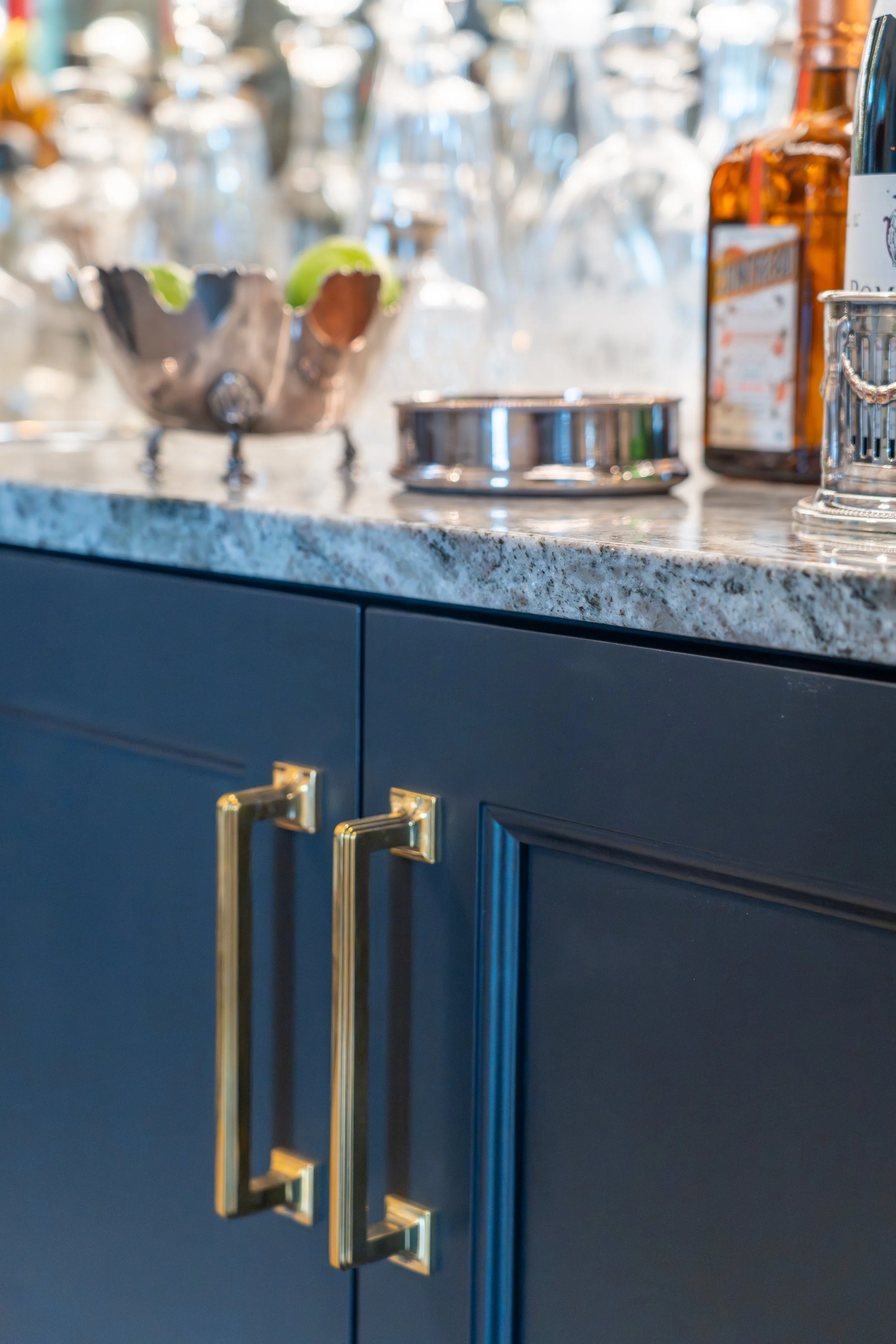 Close-up of a dark blue cabinet with gold handles, a marble countertop, and various bottles and glassware on the countertop.
