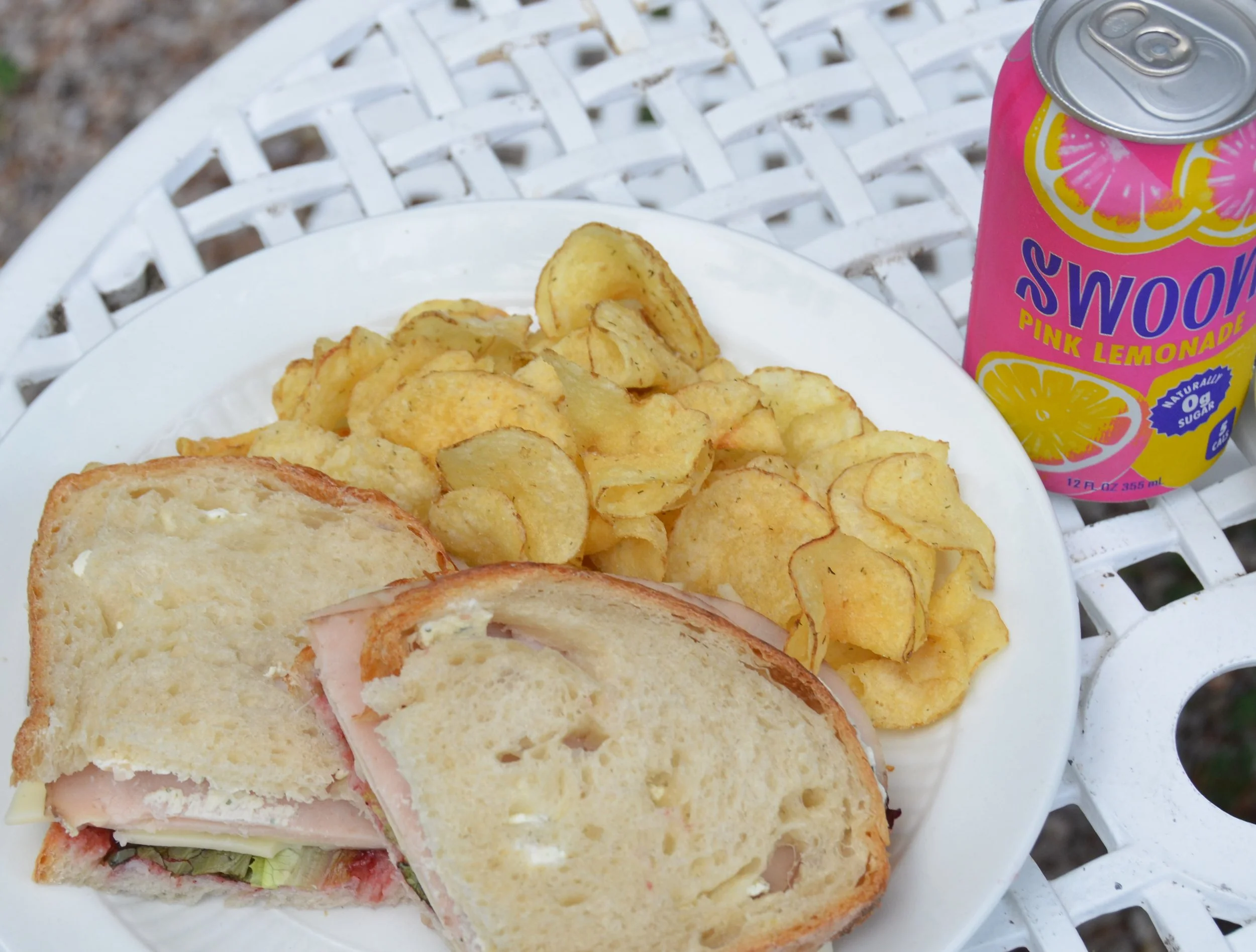Close-up of a plate with a sandwich and potato chips, with a can of pink lemonade on a white outdoor table.